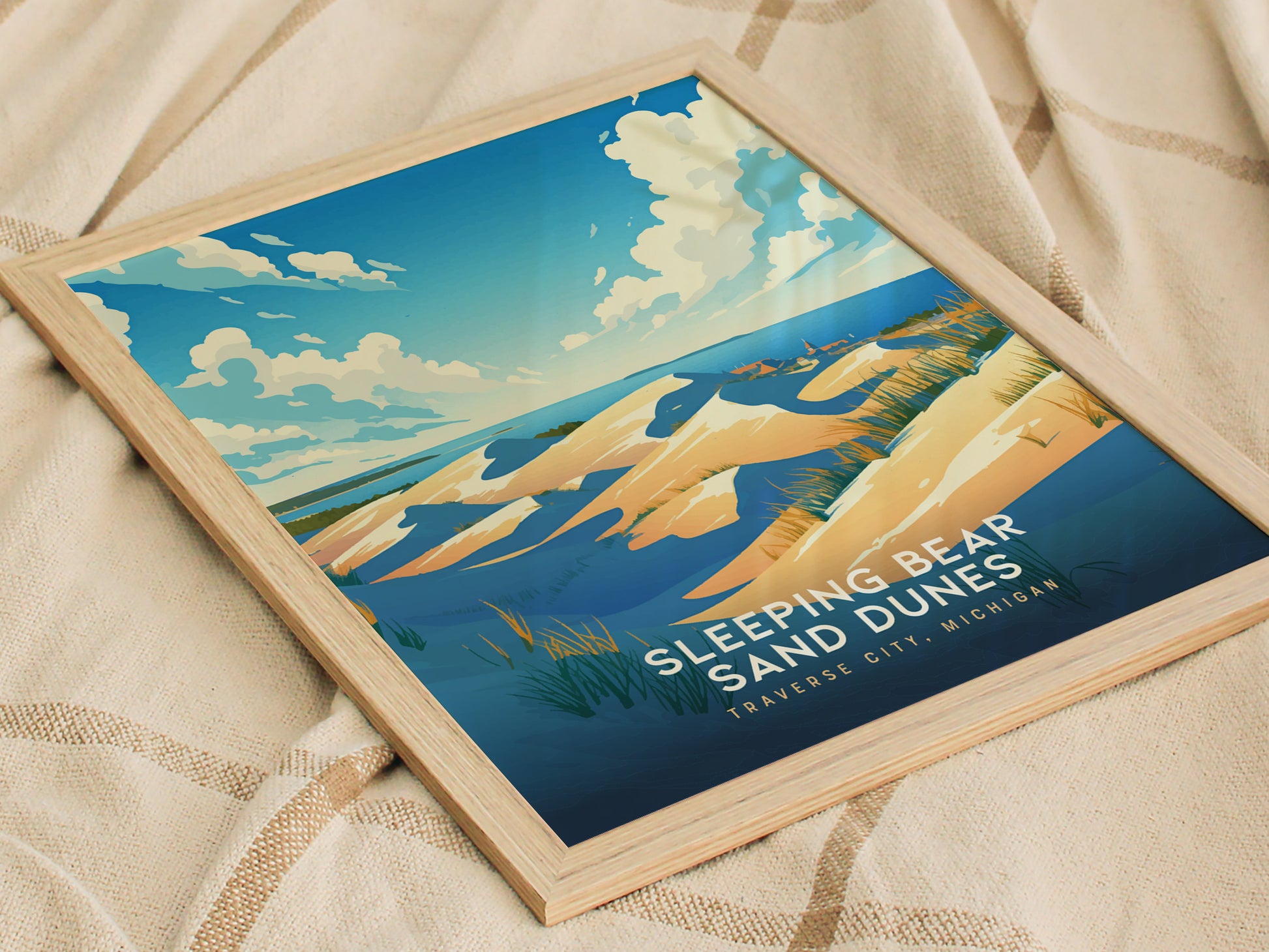 Framed artwork depicting Sleeping Bear Sand Dunes in Traverse City, Michigan, with golden sand and blue sky. The poster rests on a beige fabric background, featuring vibrant colors and scenery.