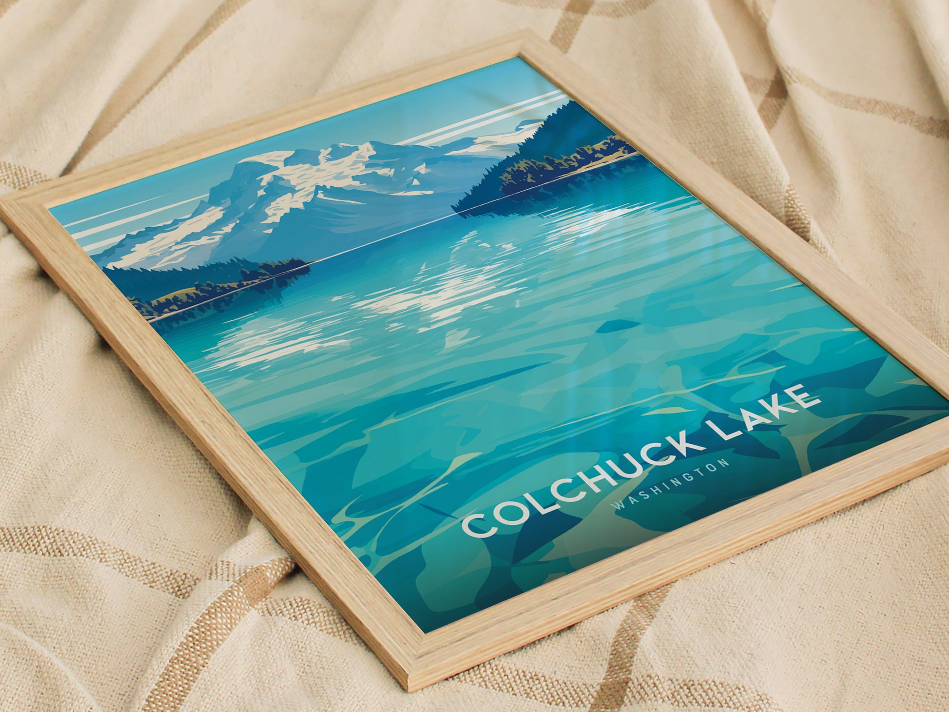 Framed poster depicting Colchuck Lake with turquoise waters and a mountainous backdrop, set on a textured beige fabric. Text reads Colchuck Lake Washington.