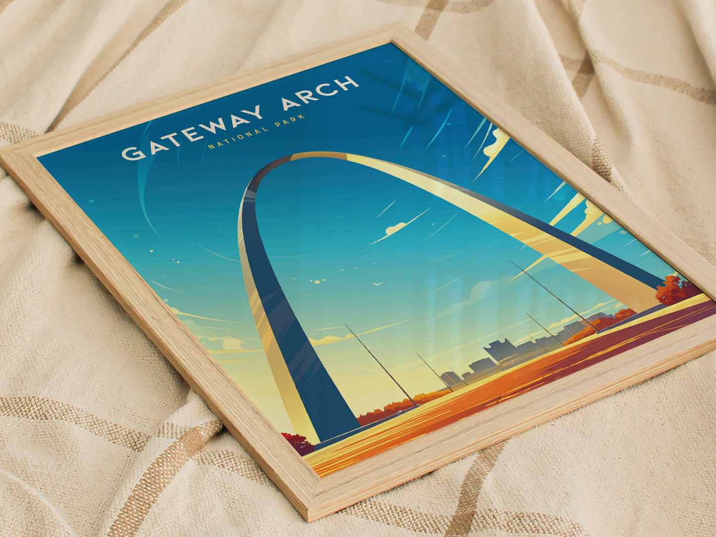 A framed poster of the Gateway Arch in St. Louis, set against a clear blue sky, lies on a beige fabric surface. The artwork features vivid colors and minimalist design.