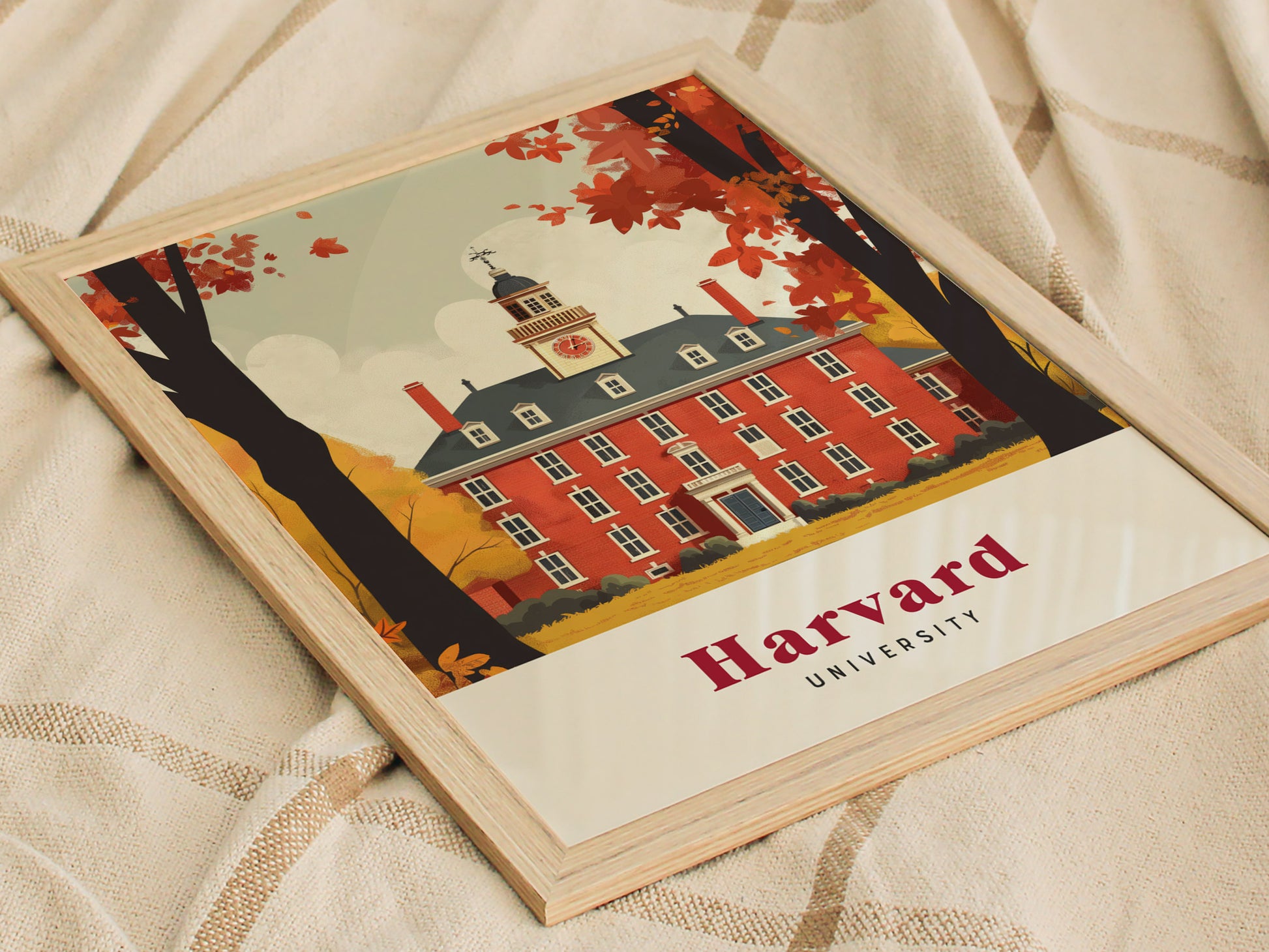 Framed illustration of a red brick Harvard University building with a clock tower, surrounded by autumn trees. The poster rests on a beige fabric surface.