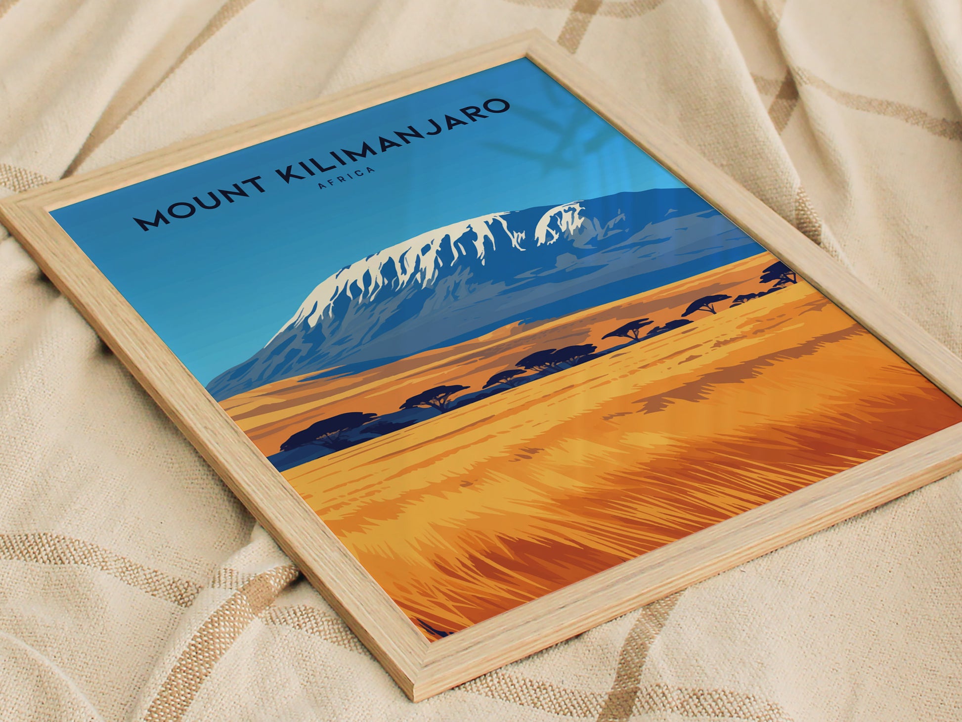 A framed poster of Mount Kilimanjaro features a vibrant landscape with a snow-capped peak and golden savanna. The artwork rests on a textured fabric surface.