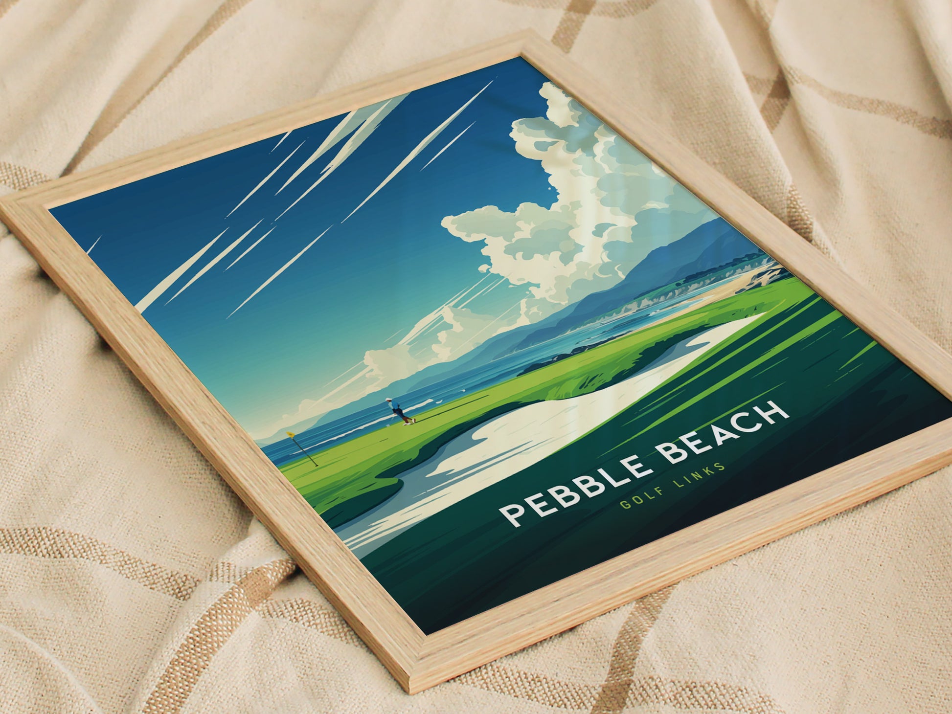 A framed Pebble Beach Golf Links poster rests on a textured beige fabric, featuring a vibrant illustration of a golfer on a lush green course with dramatic skies.