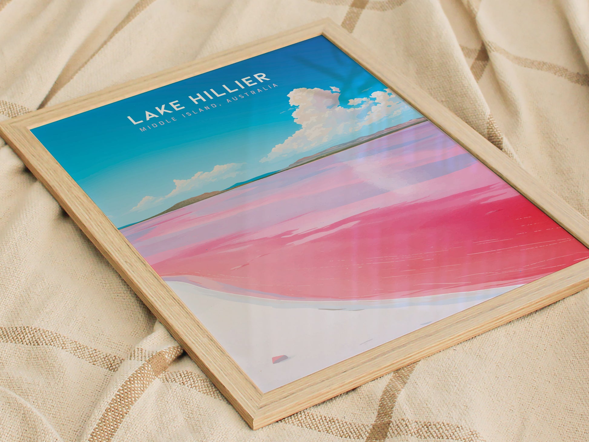 Framed poster of Lake Hillier featuring vibrant pink water and a blue sky, resting on a beige fabric. The design highlights the contrast between the lake and sky.