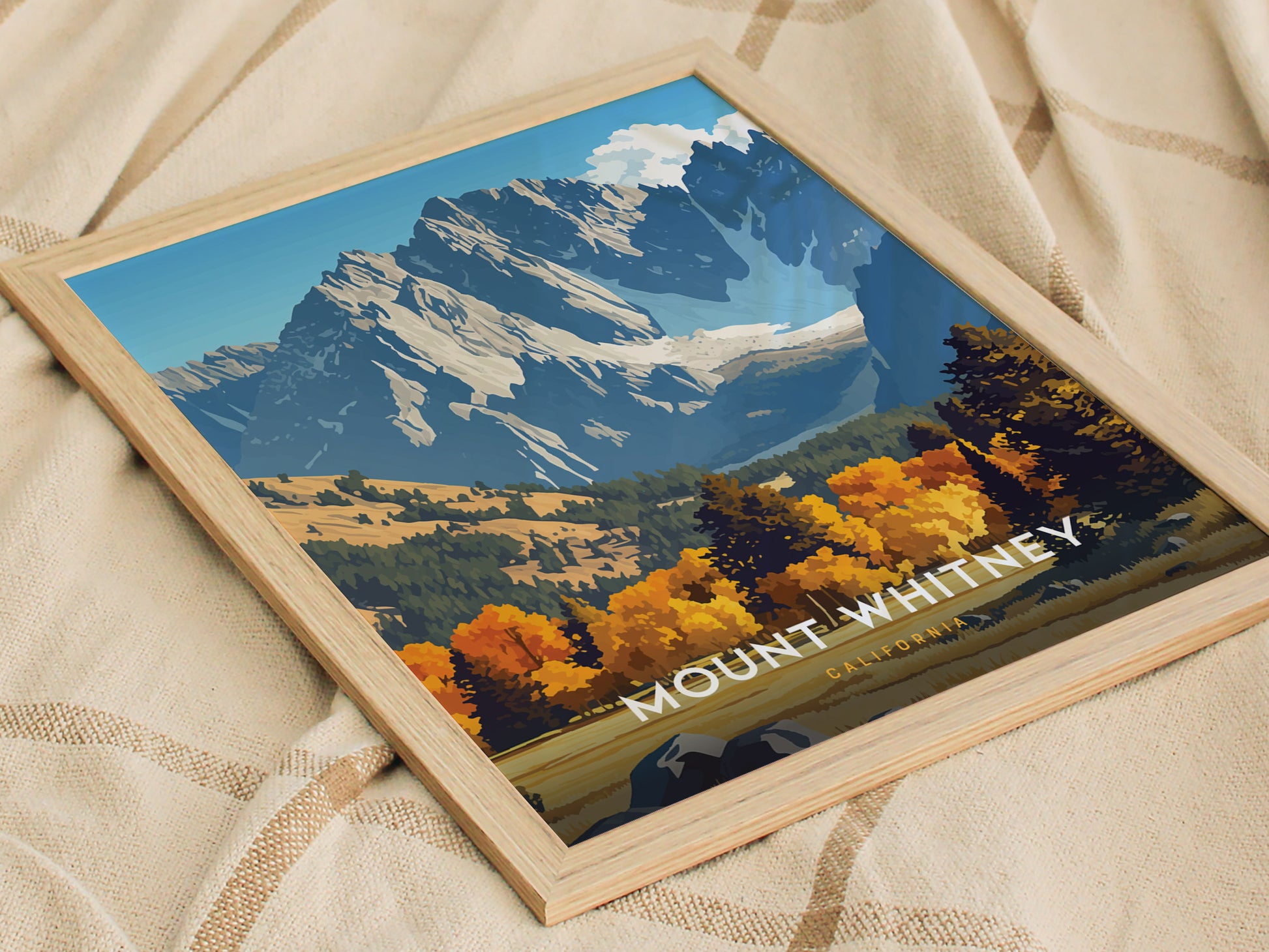 A framed poster of Mount Whitney features vibrant autumn trees and rugged granite peaks, set against a blue sky. The poster rests on a textured beige fabric.