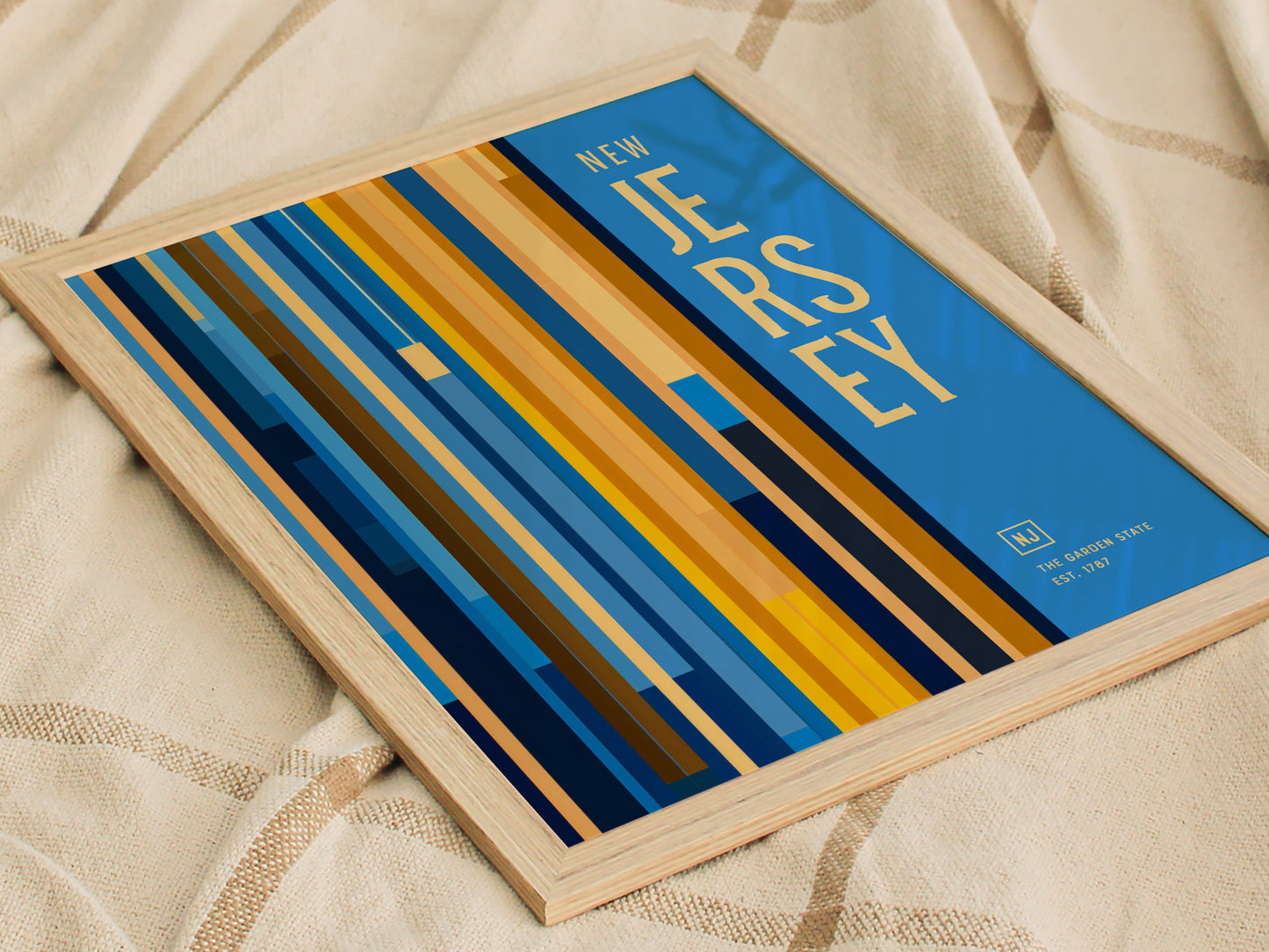Framed poster featuring bold, vertical stripes in blue, yellow, and orange with the text New Jersey on a blue background, placed on a textured fabric surface.