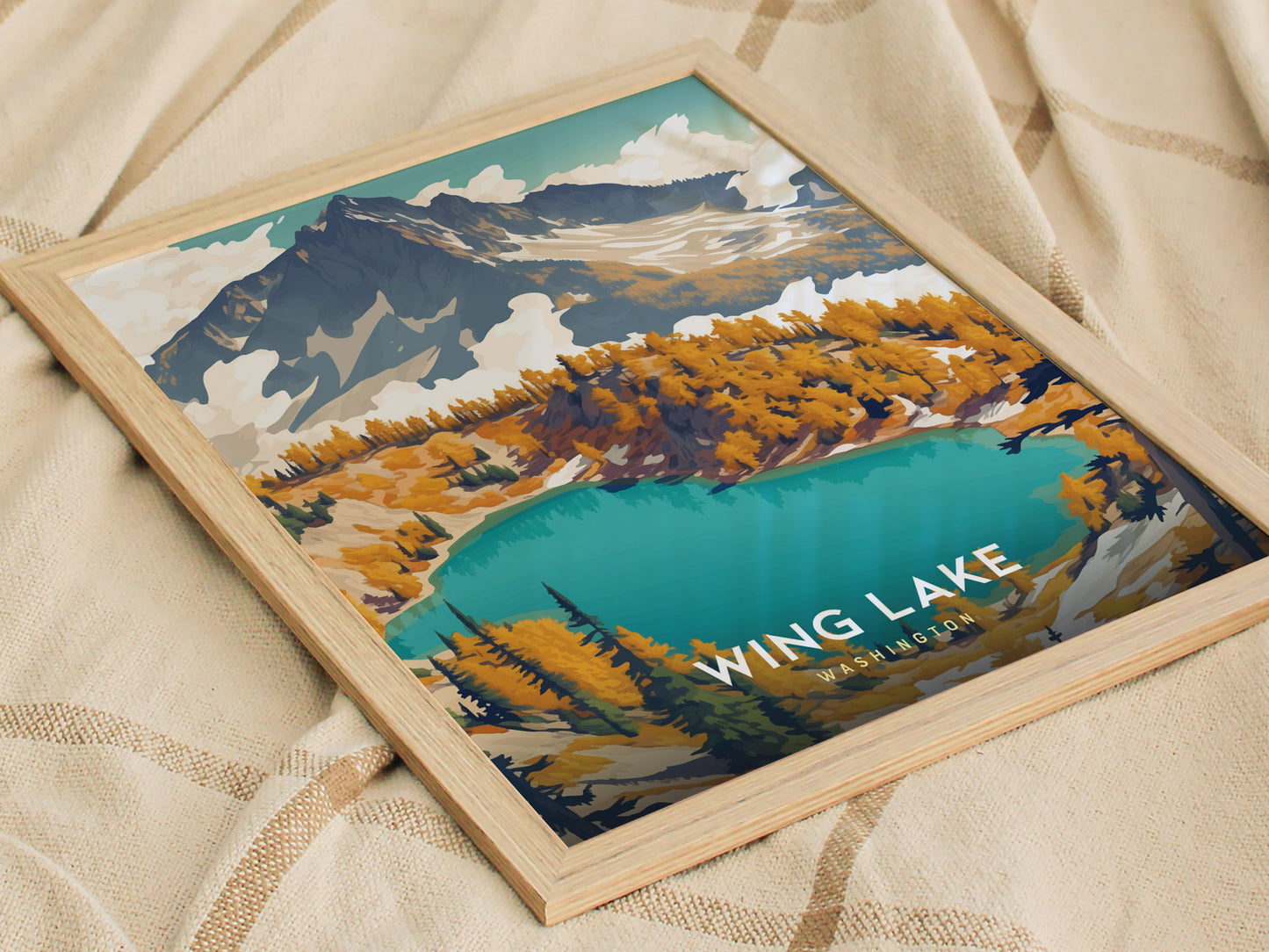 A framed poster of Wing Lake, Washington, featuring vibrant turquoise waters, surrounded by colorful autumn trees and rugged mountain peaks, rests on a beige fabric surface.