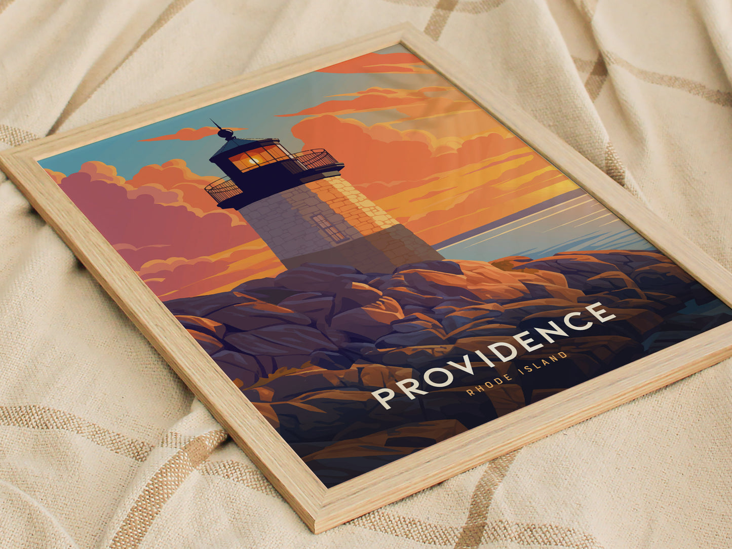 A framed poster depicting a lighthouse at sunset, with vibrant orange and purple clouds, sits on a textured beige fabric. The text reads Providence Rhode Island.