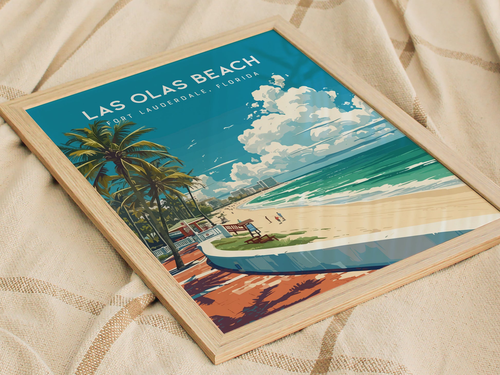 Framed Las Olas Beach poster featuring palm trees, a sandy beach, and ocean waves, resting on a textured fabric. Bright blue sky with clouds adds a tropical feel.