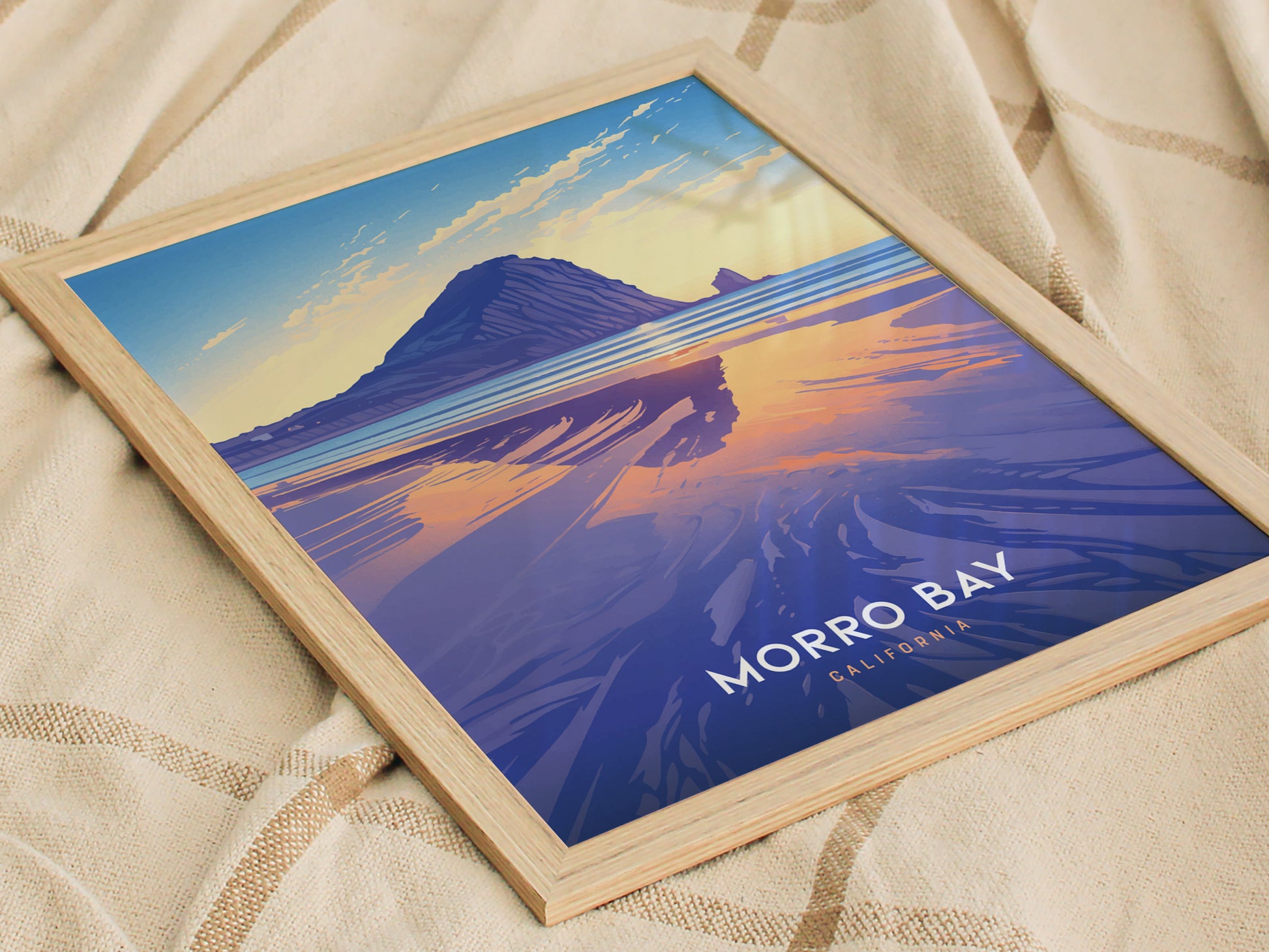 Framed poster of Morro Bay featuring a sunset view with Morro Rock reflected in the water. The artwork has vibrant blue and orange hues, placed on a textured fabric background.