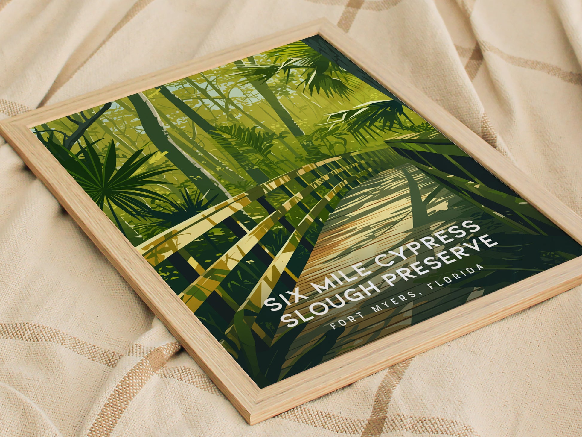 A framed poster of Six Mile Cypress Slough Preserve rests on a textured fabric, depicting a wooden boardwalk surrounded by lush green foliage.