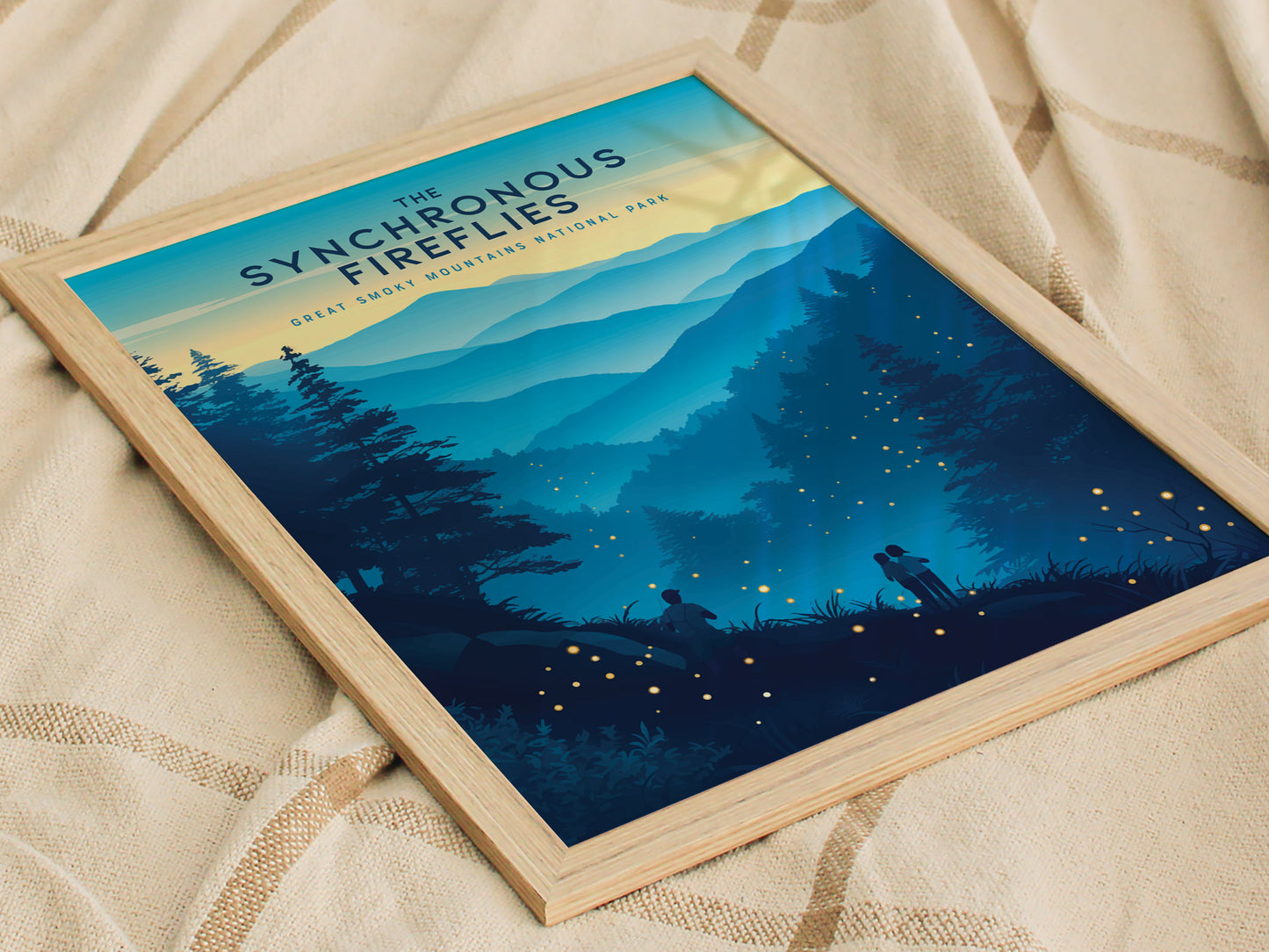 A framed poster titled The Synchronous Fireflies depicts a serene landscape of the Great Smoky Mountains with silhouettes of figures watching glowing fireflies under a twilight sky. The poster rests on a soft, textured blanket.
