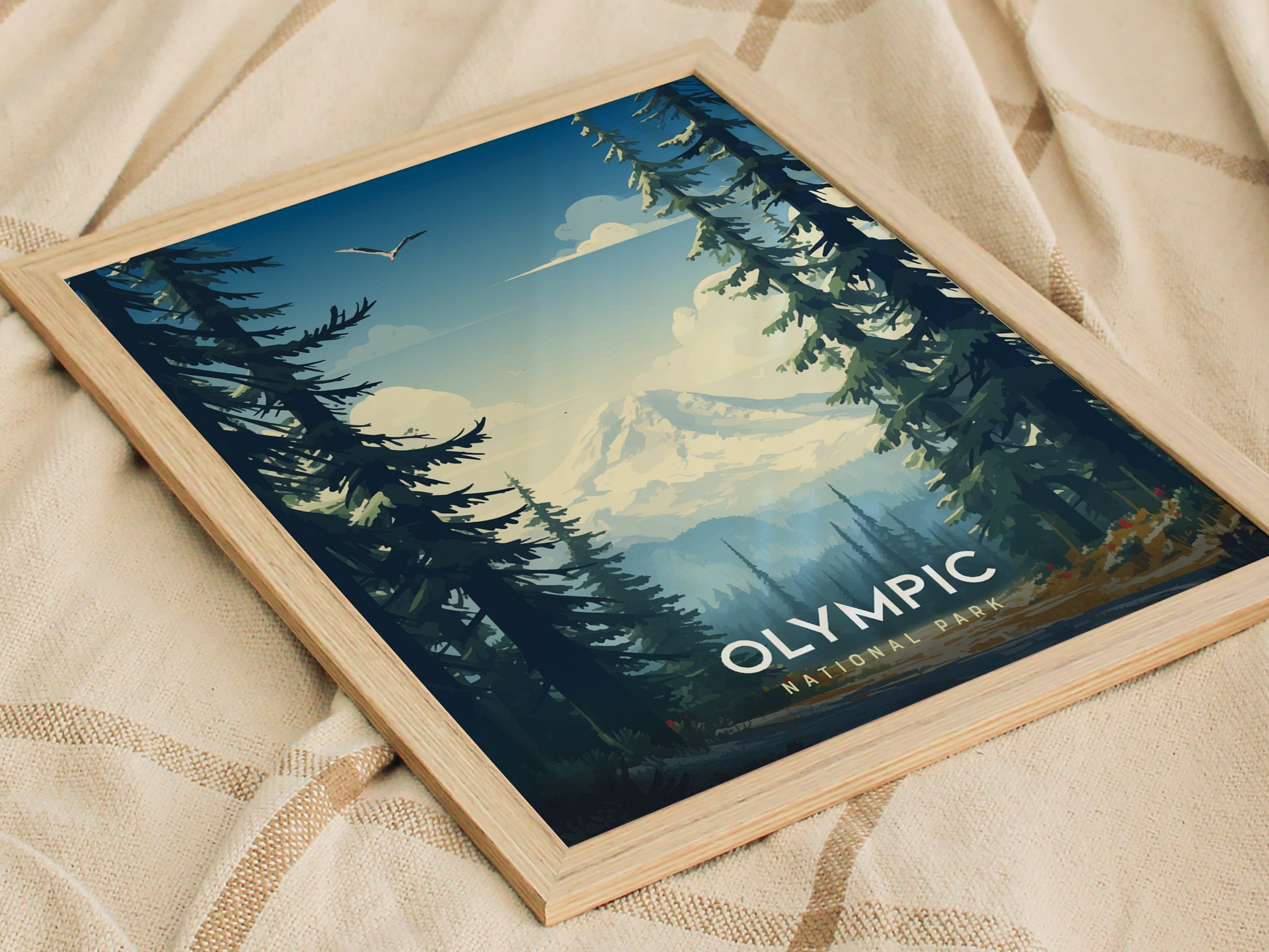 Framed Olympic National Park poster featuring a scenic view of pine trees, distant mountains, and a bird soaring in the sky, placed on a textured fabric surface.