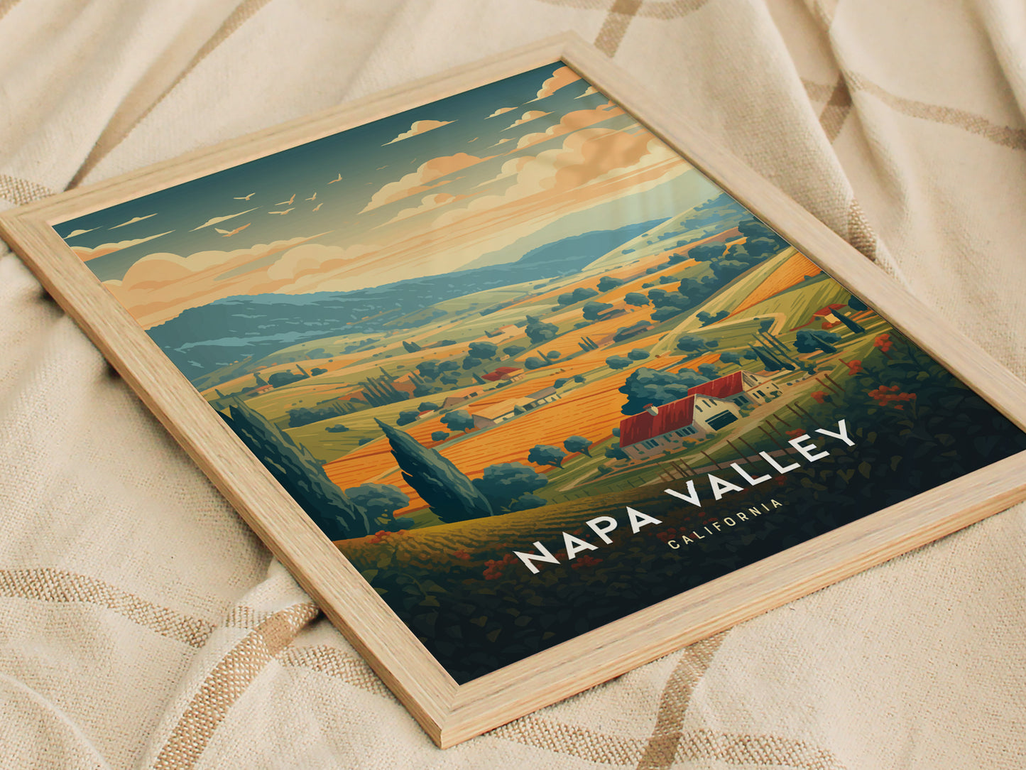 Framed Napa Valley travel poster featuring rolling vineyards, tree-lined hills, and a warm sunset sky. The artwork is set against a soft, textured fabric background.