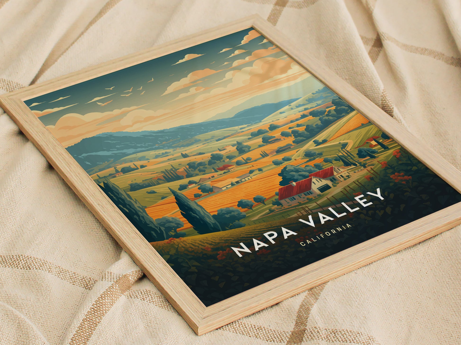 Framed Napa Valley travel poster featuring rolling vineyards, tree-lined hills, and a warm sunset sky. The artwork is set against a soft, textured fabric background.