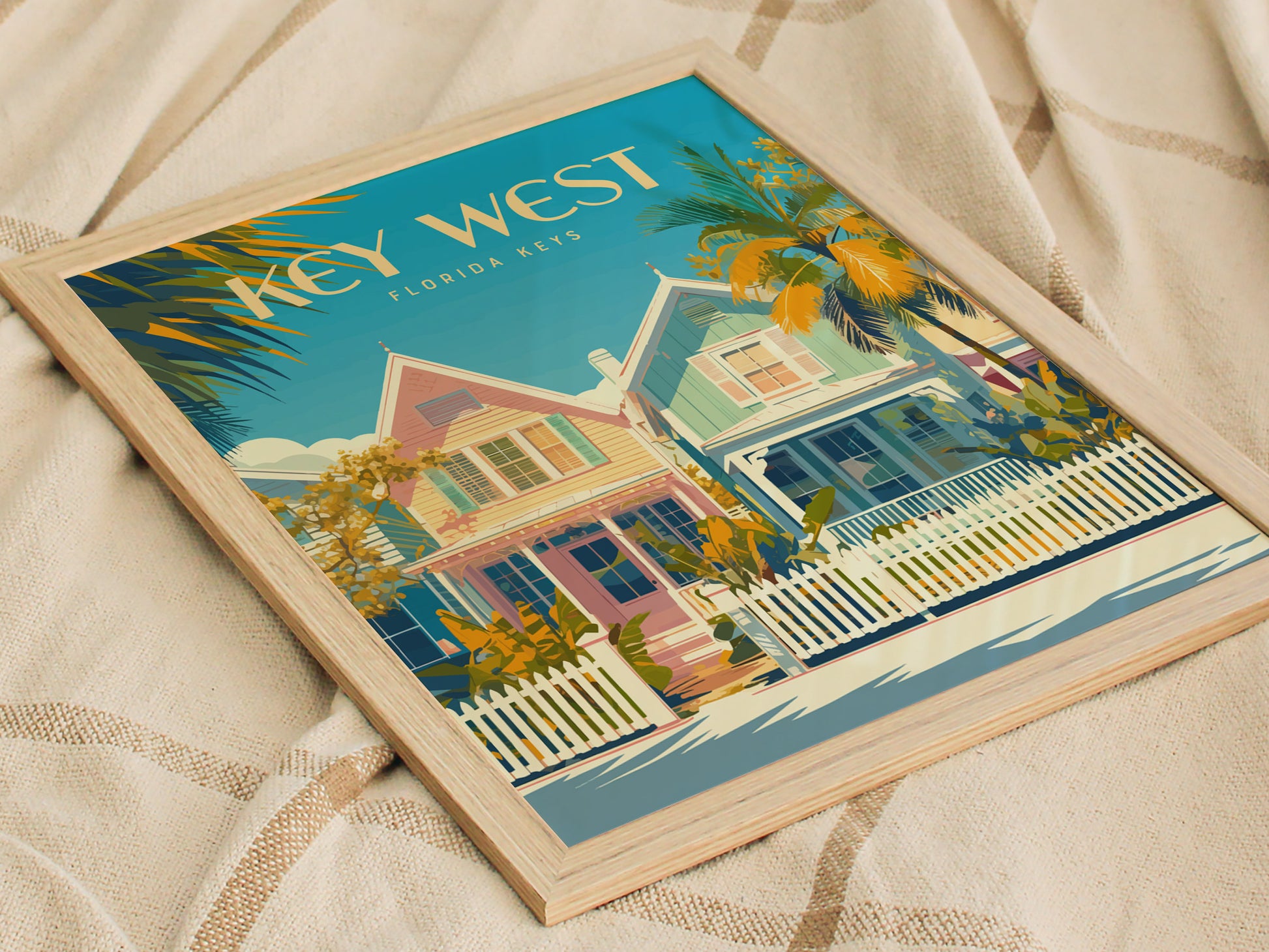Framed Key West poster featuring pastel-colored houses, palm trees, and a white picket fence. The artwork rests on a beige, patterned fabric surface, capturing a tropical, relaxed vibe.