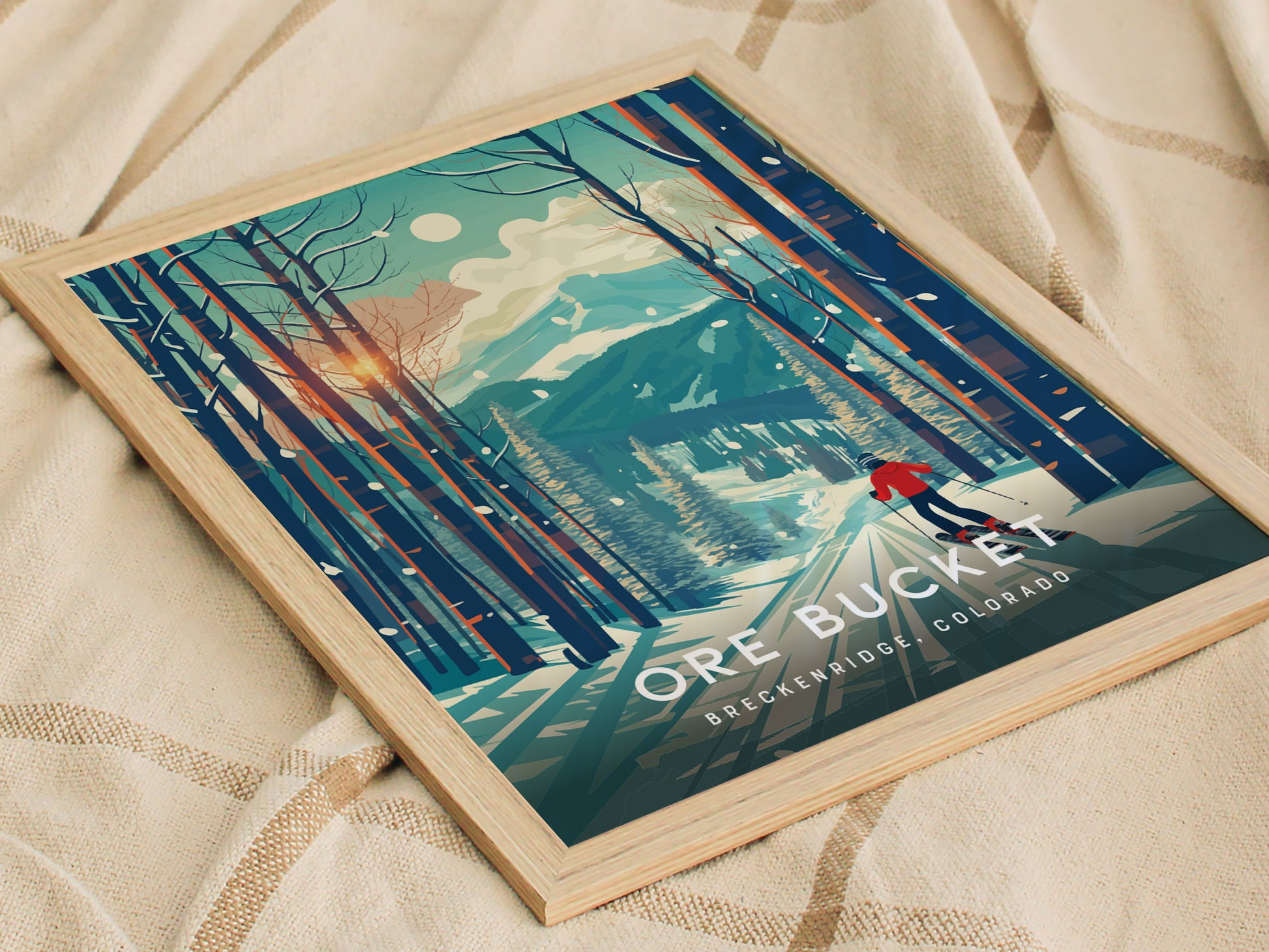 Framed poster featuring a stylized winter scene with a skier on a mountain trail surrounded by tall trees, titled Ore Bucket Breckenridge, Colorado, resting on a beige fabric.