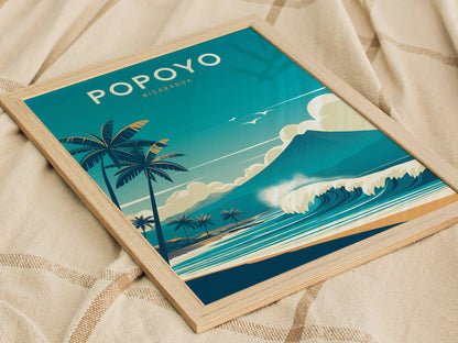 Framed poster featuring a stylized illustration of Popoyo, Nicaragua, with palm trees, waves, and a mountain against a blue sky, resting on a beige fabric surface.