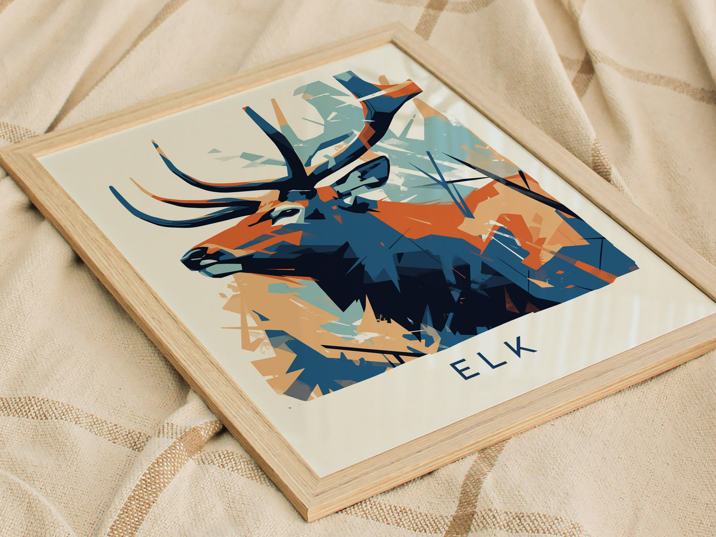 A modern abstract print featuring a stylized elk with bold geometric shapes in shades of blue and orange. The artwork is framed and positioned on a textured, neutral-colored fabric background, showcasing its vibrant design.