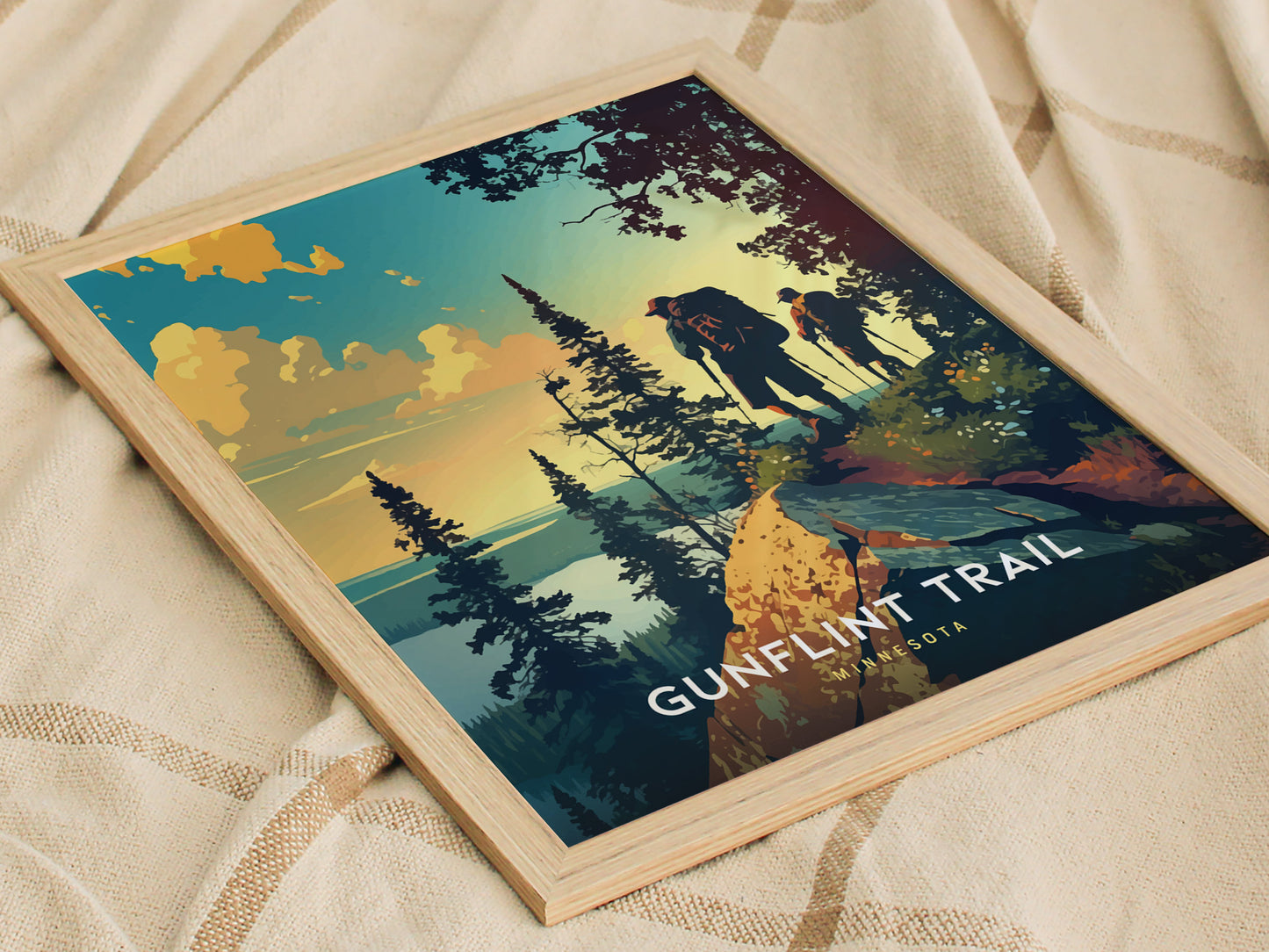 Framed poster depicting two hikers on a forested trail with a vibrant sunset in the background, labeled Gunflint Trail Minnesota. The artwork rests on a textured fabric surface.