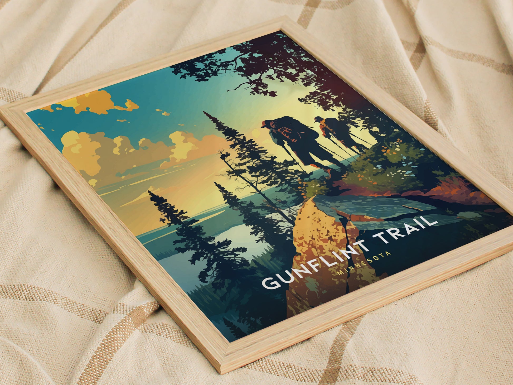 Framed poster depicting two hikers on a forested trail with a vibrant sunset in the background, labeled Gunflint Trail Minnesota. The artwork rests on a textured fabric surface.