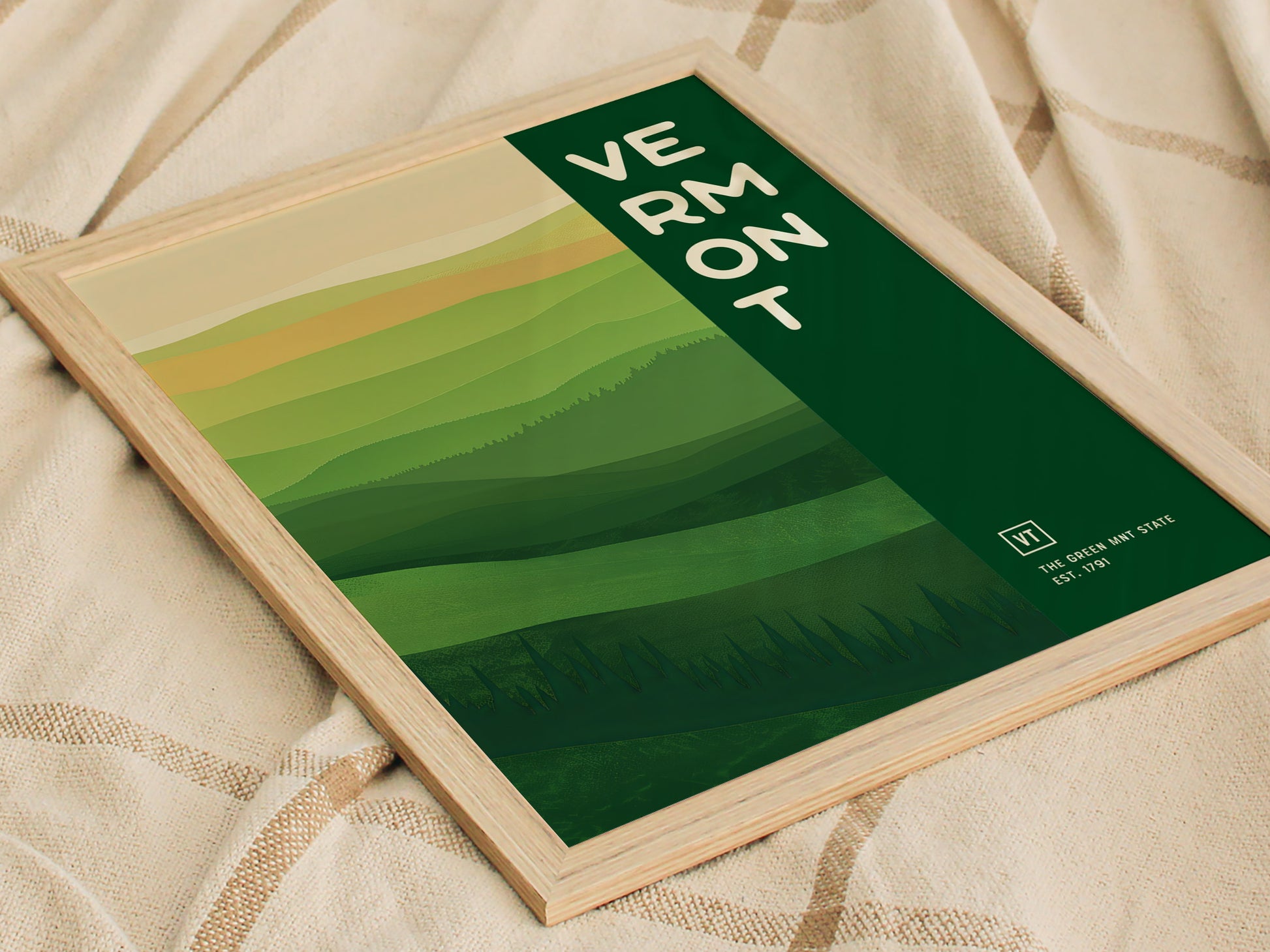 A framed Vermont poster featuring abstract rolling green hills and the text Vermont in bold white letters. The artwork rests on a textured beige fabric.