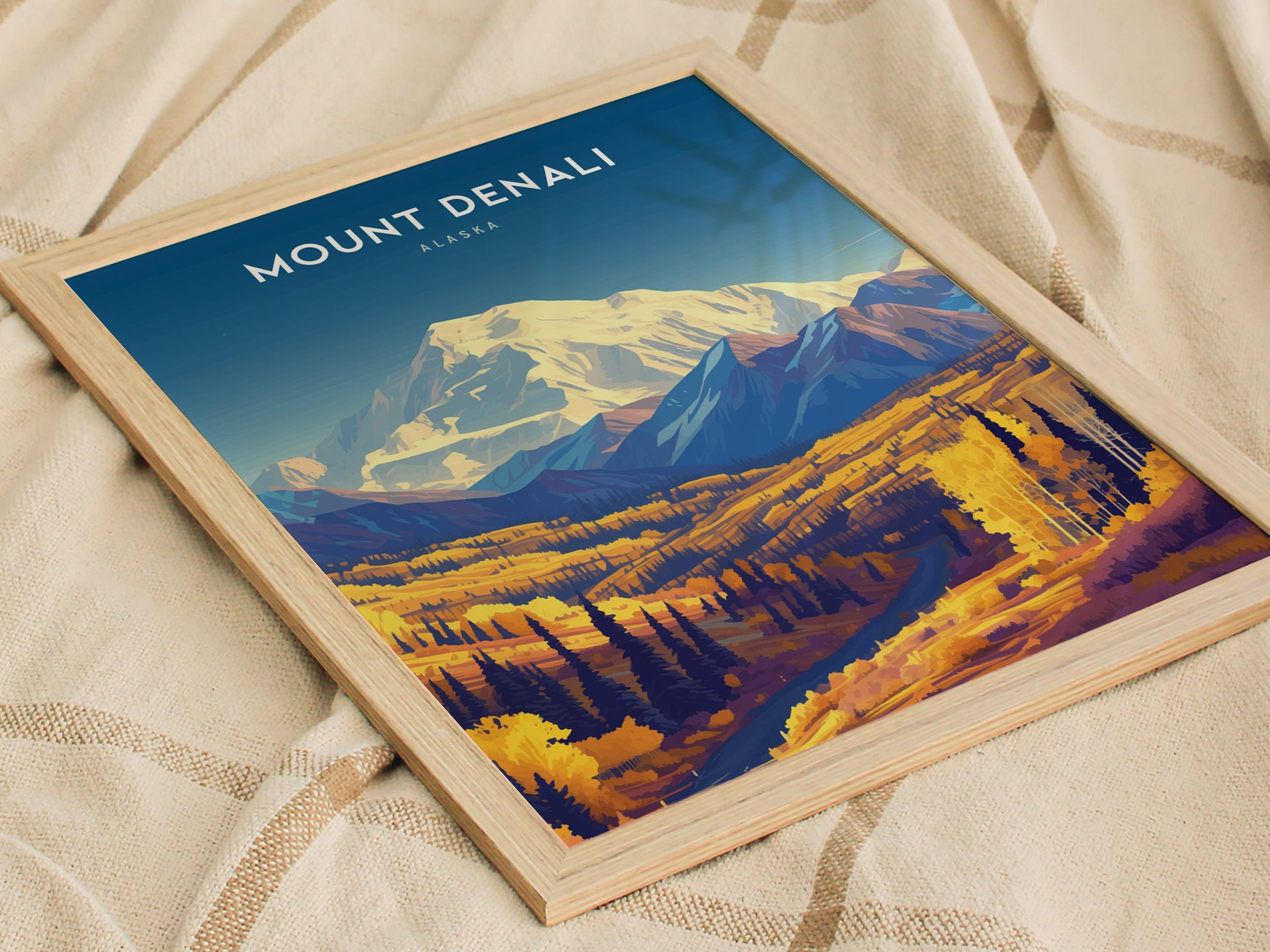 Framed Mount Denali poster featuring a vibrant illustration of the Alaskan landscape with golden foliage and a snowy peak. The poster is set against a soft, textured fabric background.