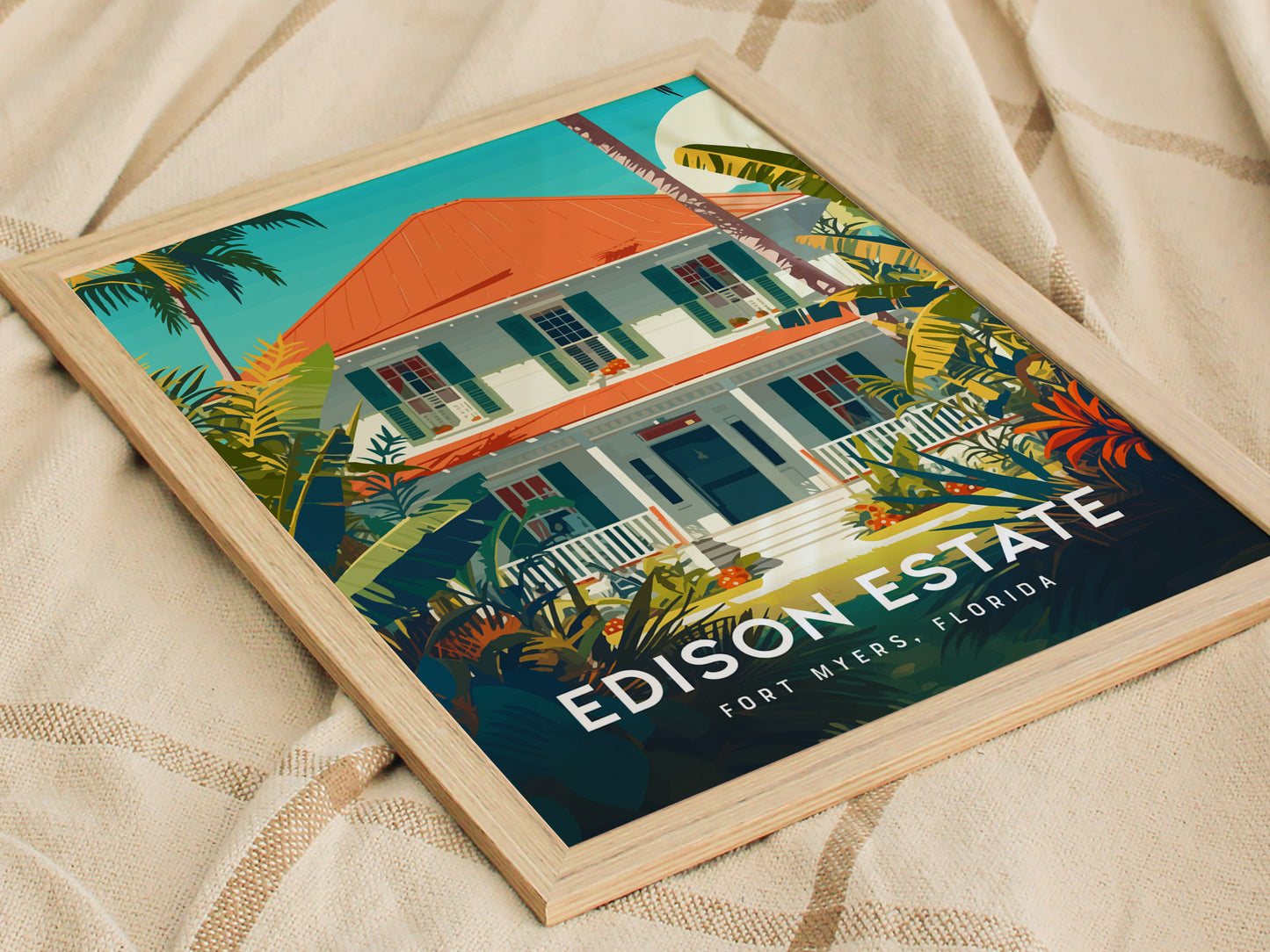 Framed poster featuring a colorful illustration of a house with a red roof, surrounded by lush tropical plants, titled Edison Estate, Fort Myers, Florida, resting on a beige fabric.