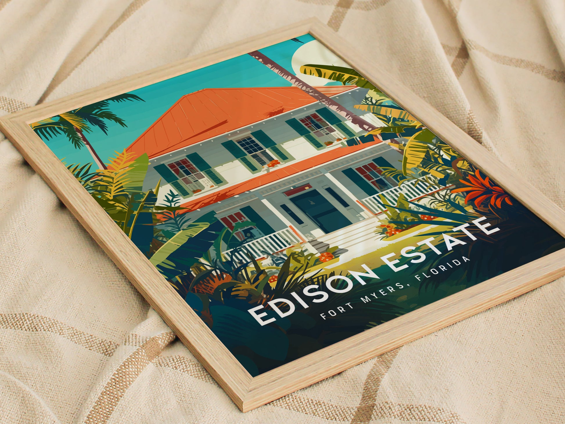 Framed poster featuring a colorful illustration of a house with a red roof, surrounded by lush tropical plants, titled Edison Estate, Fort Myers, Florida, resting on a beige fabric.
