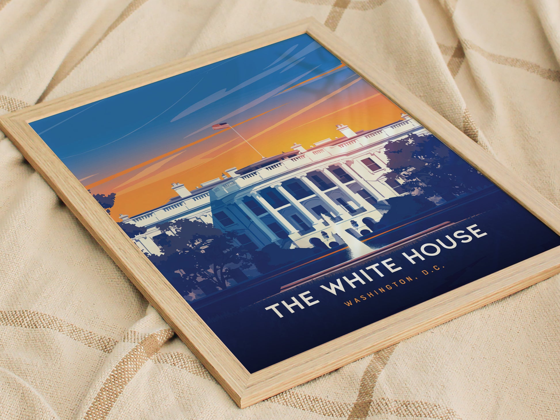 A framed poster of the White House with a vibrant sunset background, featuring bold colors and minimalist design, lies on a textured fabric surface.