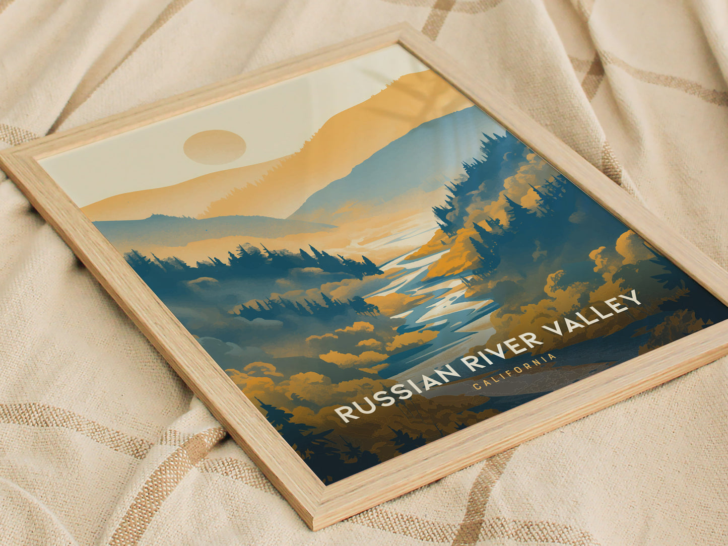 A framed poster titled Russian River Valley, California depicts a scenic landscape with rolling hills, a river, and a sun setting behind the mountains, placed on a textured fabric backdrop.