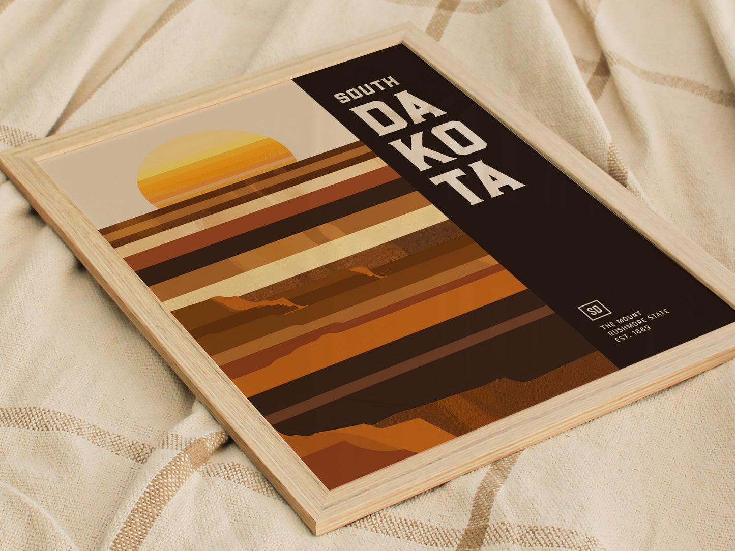 Framed South Dakota poster featuring minimalist geometric art with earthy tones and horizontal stripes, depicting a setting sun and abstract landscape, resting on a textured fabric background.