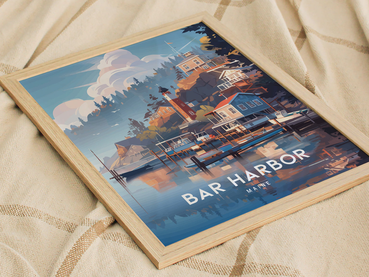 Framed poster of Bar Harbor, Maine, depicting colorful coastal houses and boats on a calm waterfront, set against a backdrop of trees and a cloudy sky, resting on a fabric.