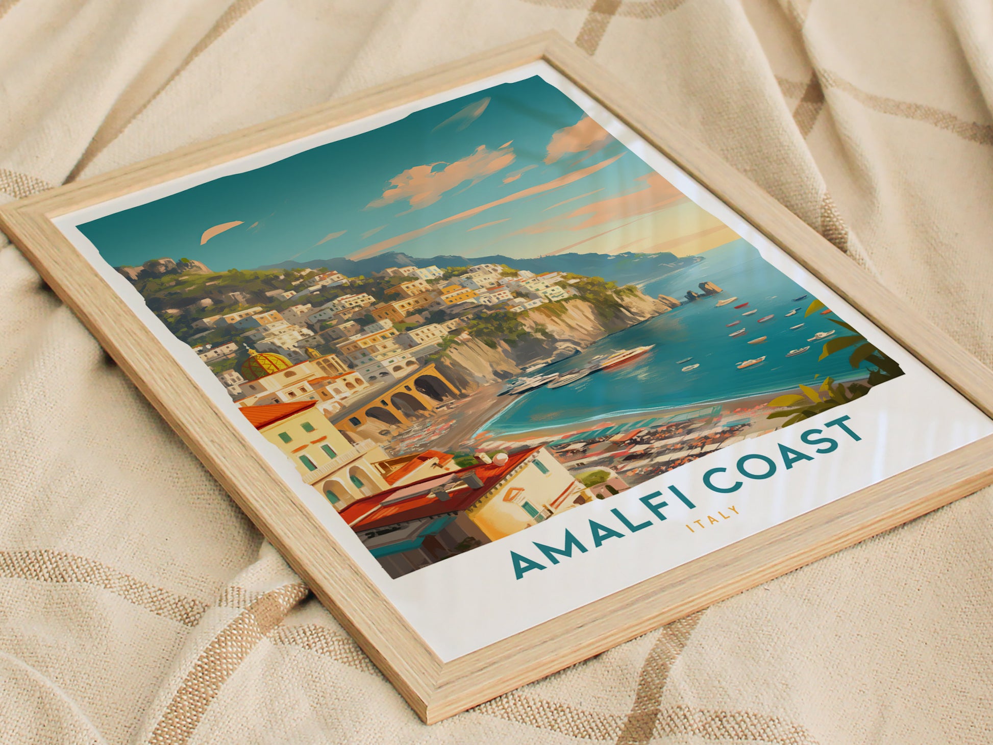 Framed artwork depicting the Amalfi Coast with colorful buildings on cliffs above a turquoise sea, set on a beige fabric background. The text reads Amalfi Coast Italy.