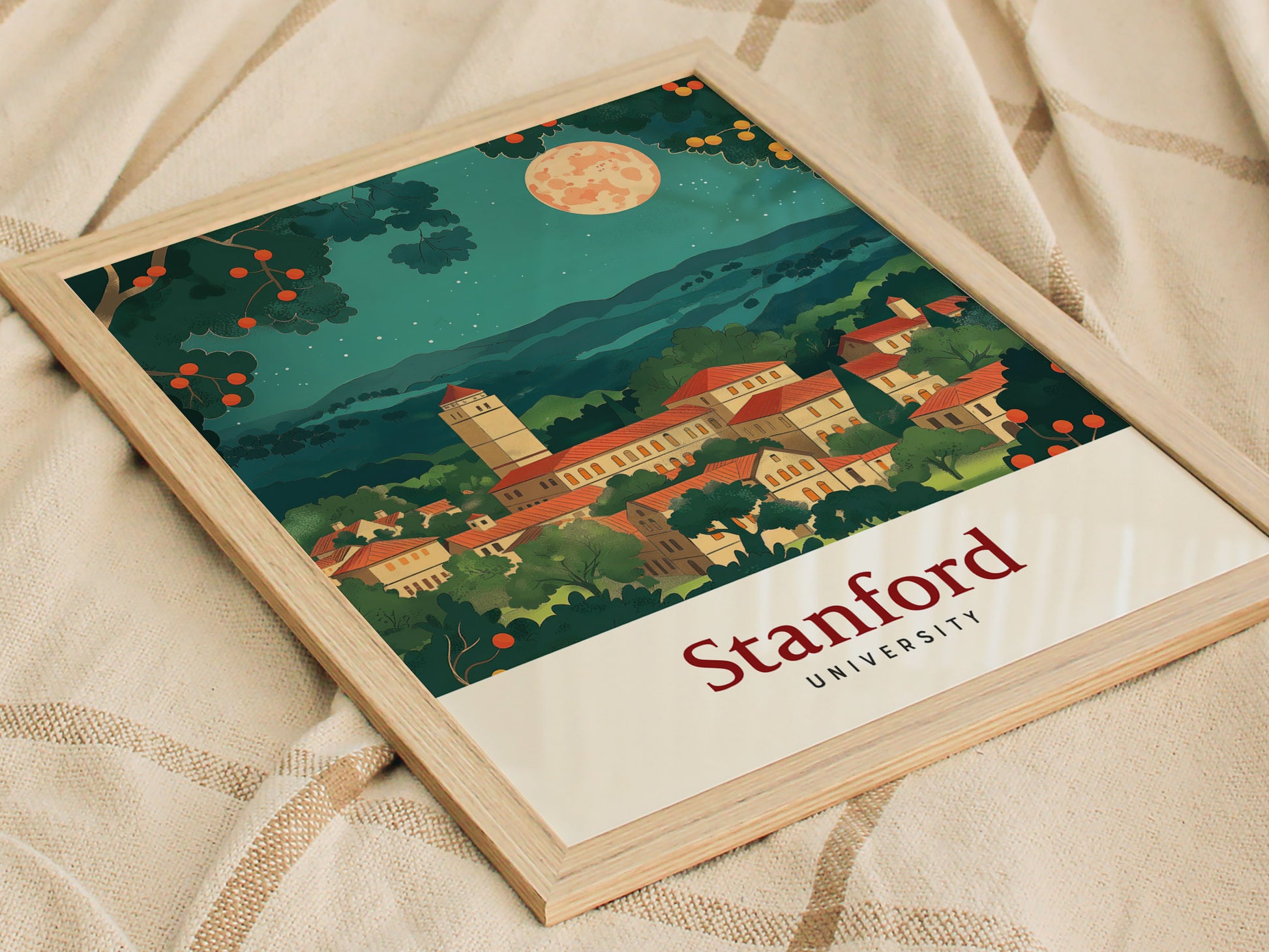 A framed Stanford University poster features a vintage-style illustration of campus buildings under a starry sky with a full moon, set on a textured fabric surface.
