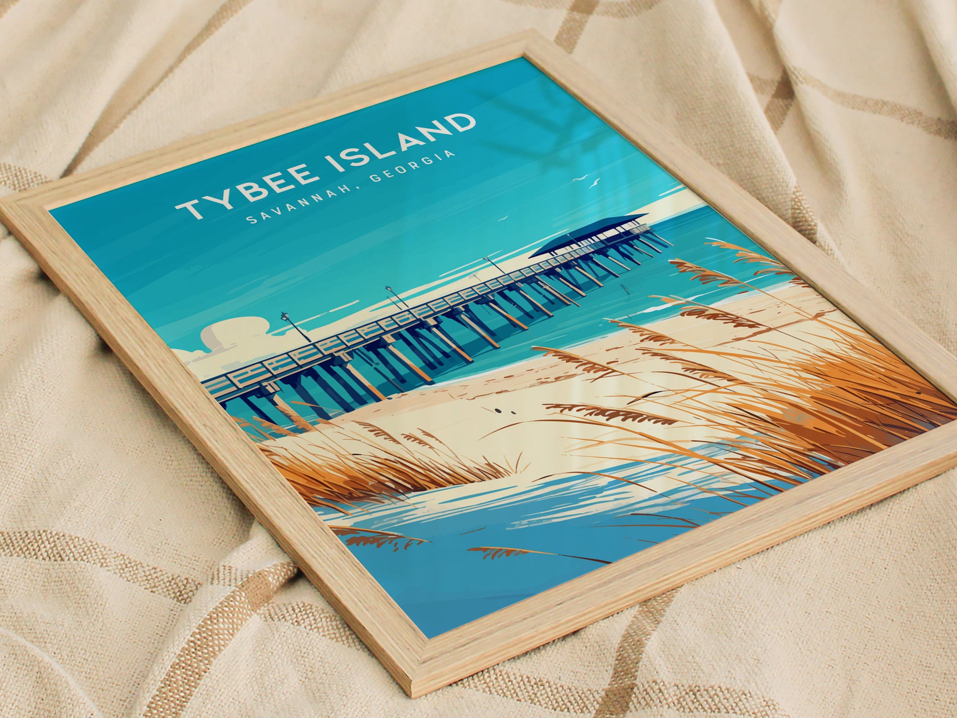 A framed poster of Tybee Island, Savannah, Georgia, featuring a vibrant illustration of a fishing pier, sandy dunes, and sea oats, rests on a textured fabric background.