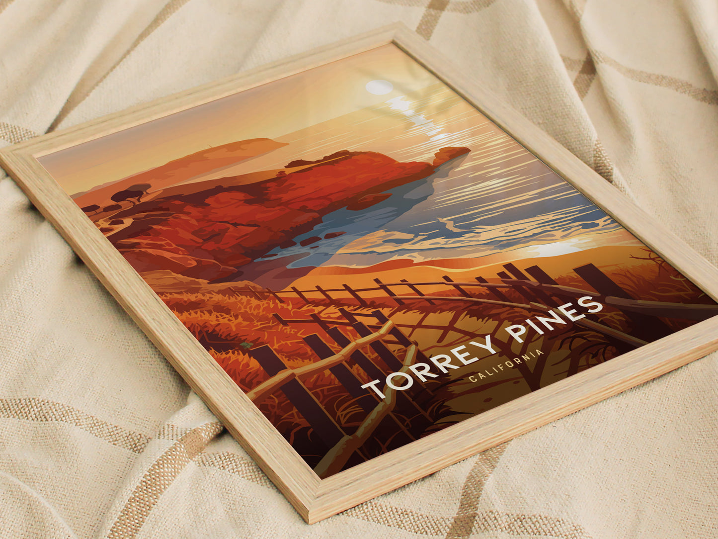 A framed poster featuring an illustration of Torrey Pines, California, with vibrant orange cliffs, a winding trail, and the ocean under a setting sun, resting on a beige fabric.