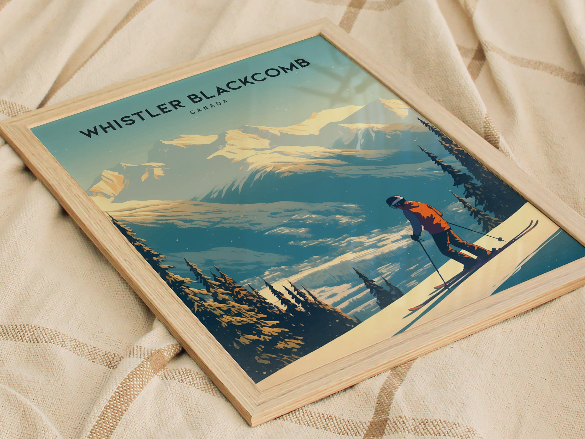 Framed poster of Whistler Blackcomb, featuring a skier in an orange jacket against snow-covered mountains and pine trees, laid on a textured beige fabric.