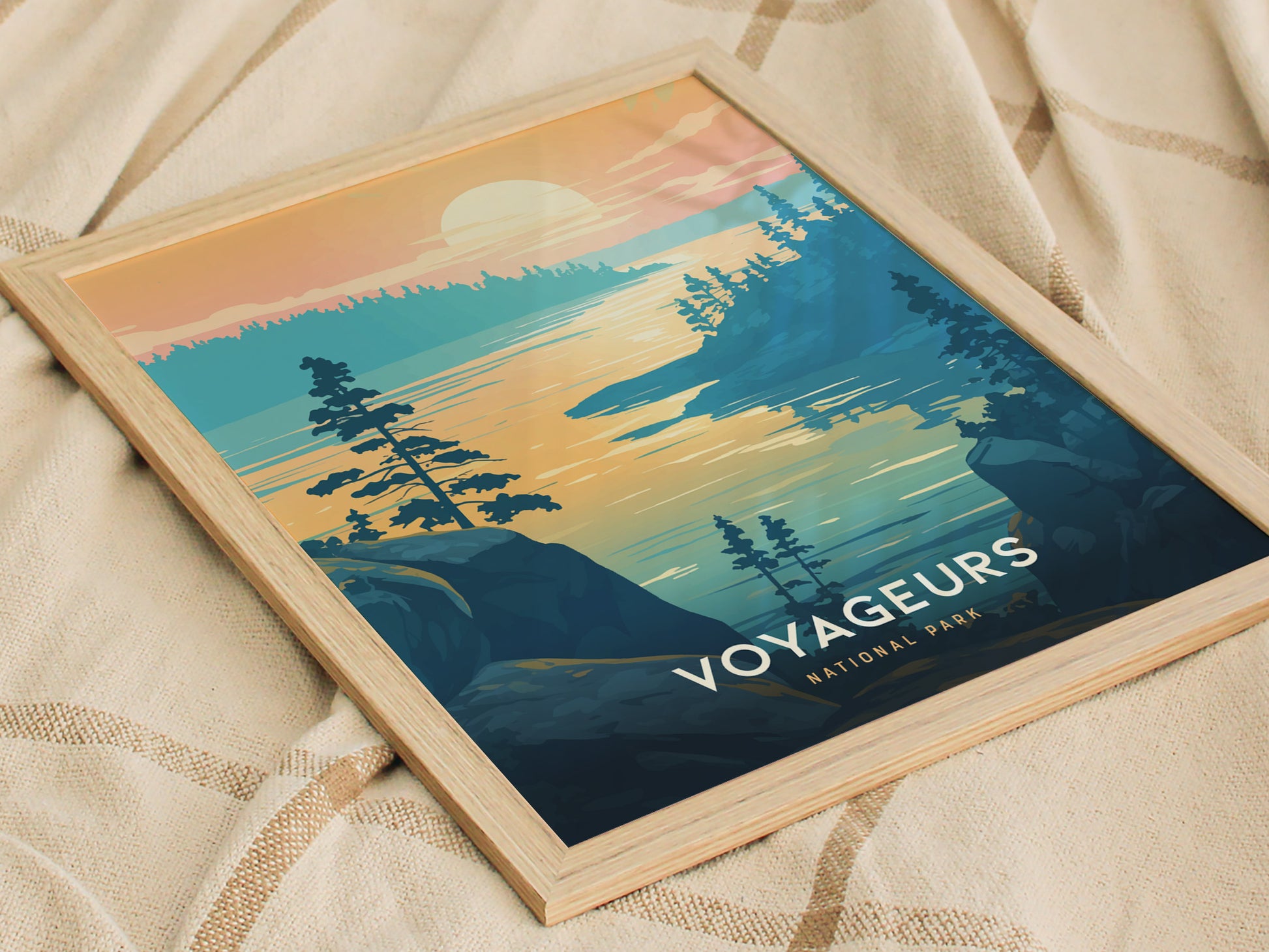Framed artwork of Voyageurs National Park features a serene lake scene with trees silhouetted against a colorful sunset. The poster rests on a soft, textured beige fabric.