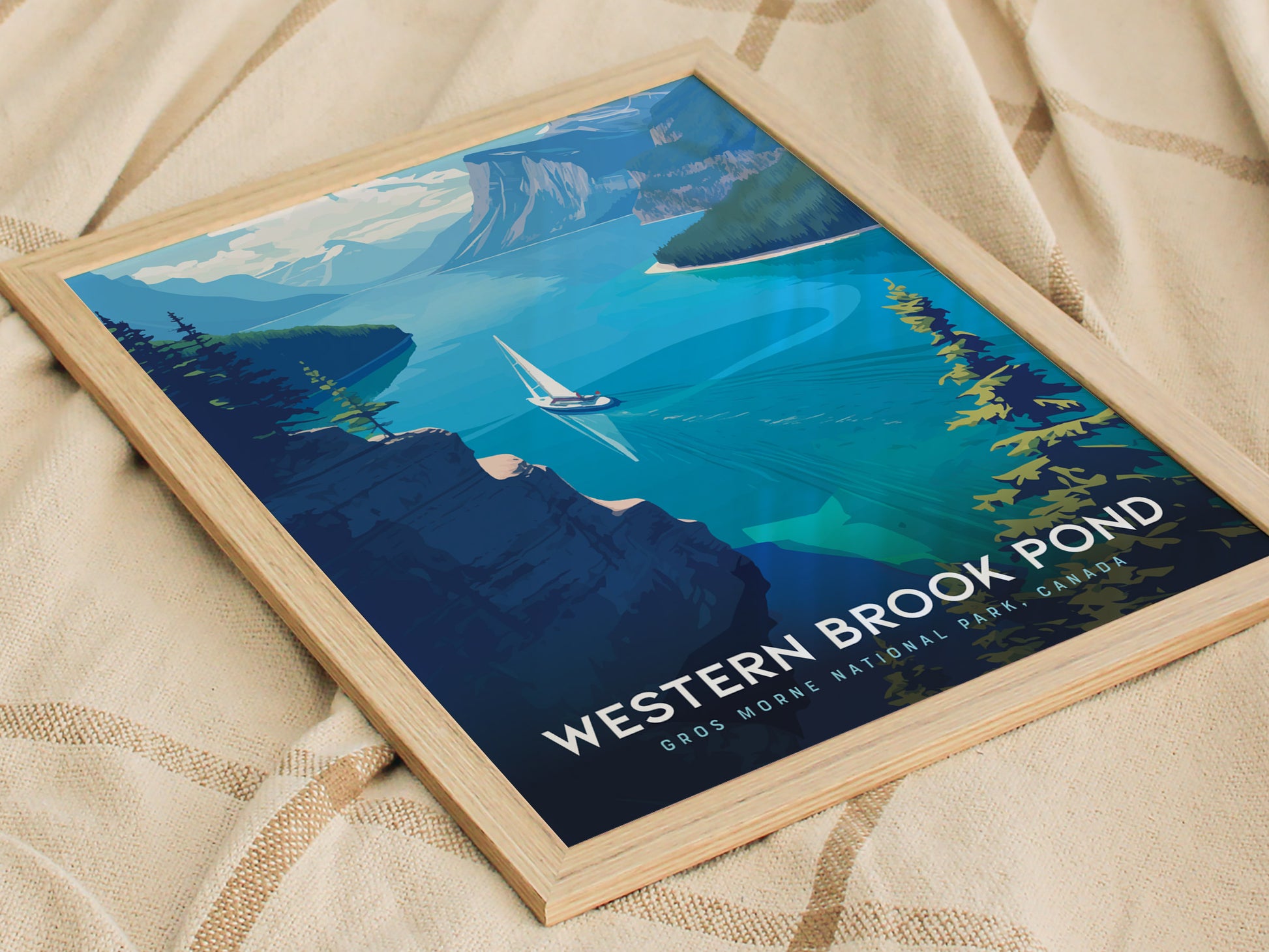 A framed poster of Western Brook Pond shows a sailboat gliding on turquoise waters surrounded by dramatic cliffs and lush greenery, lying on a textured beige fabric.