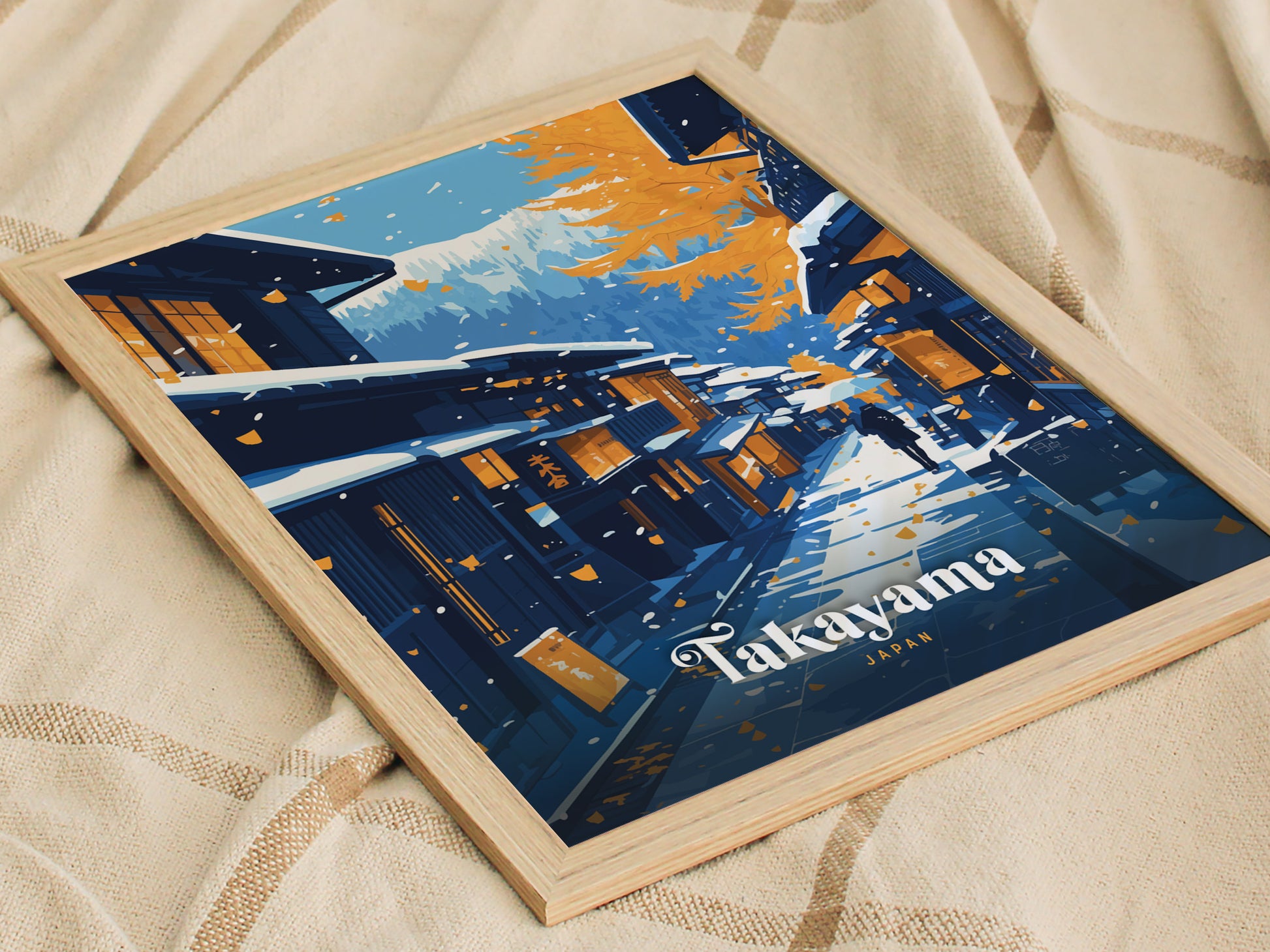 Framed artwork of Takayama, Japan, featuring stylized streets with autumn leaves and traditional buildings under a snowy mountain backdrop, resting on a textured fabric.