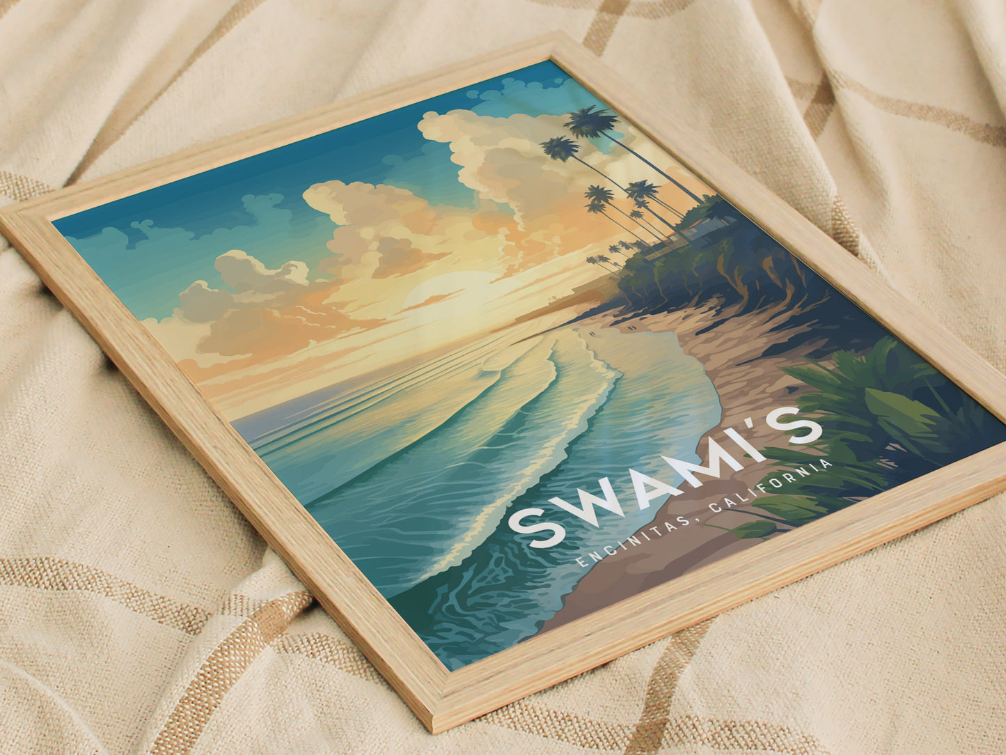 Framed artwork of Swamis Beach, Encinitas, California, depicting a sunset view with waves, cliffs, and palm trees. The poster rests on a beige fabric surface.