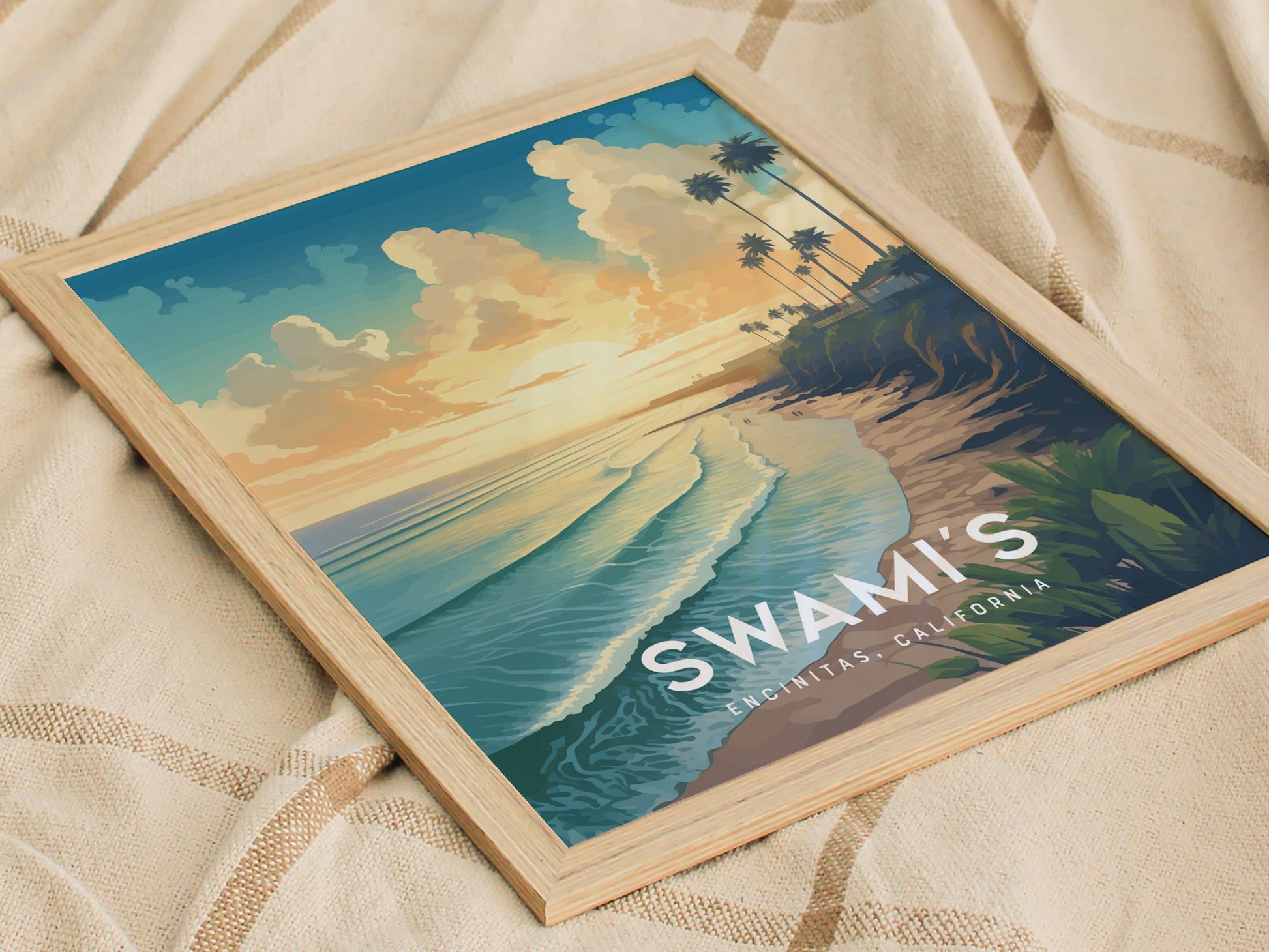Framed artwork of Swamis Beach, Encinitas, California, depicting a sunset view with waves, cliffs, and palm trees. The poster rests on a beige fabric surface.