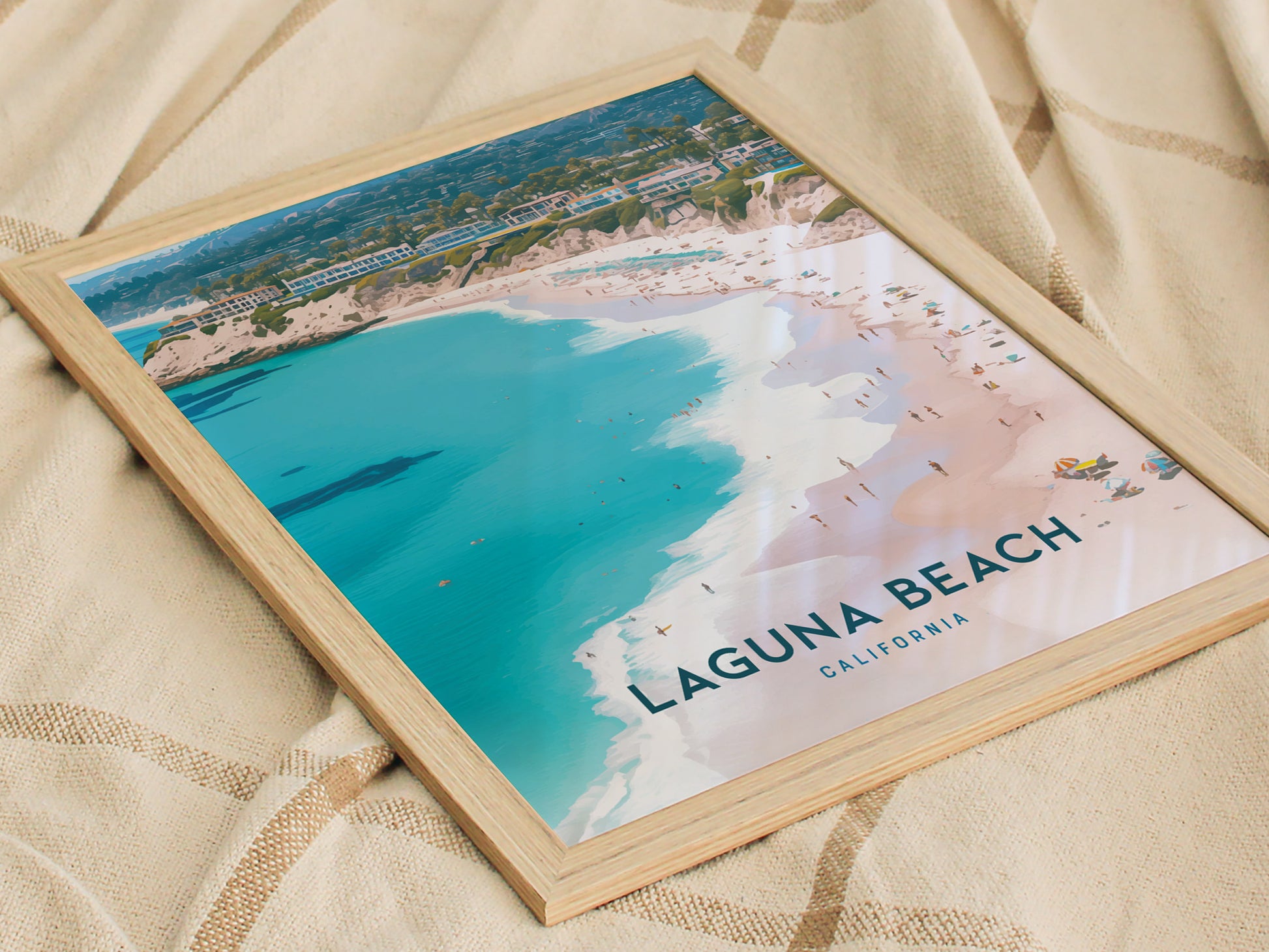 Framed poster of Laguna Beach, California, showcasing a vibrant aerial view of turquoise waters, sandy shores, and distant cliffs, placed on a textured beige fabric.