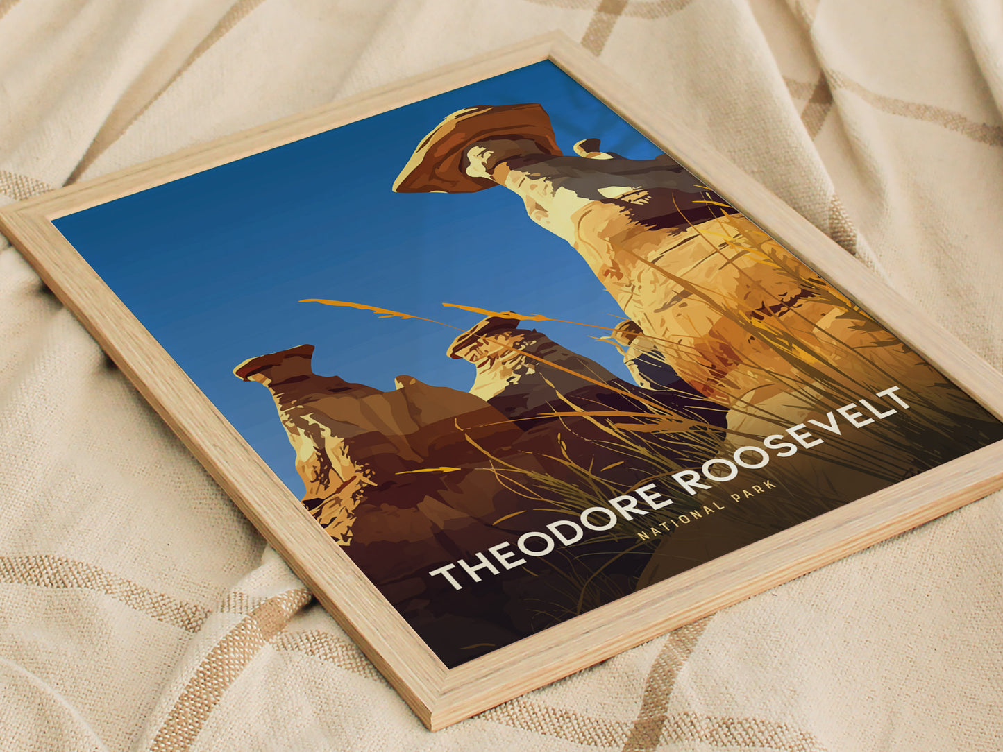 A framed poster featuring stylized hoodoos against a clear blue sky, with Theodore Roosevelt National Park text, rests on a textured beige fabric.