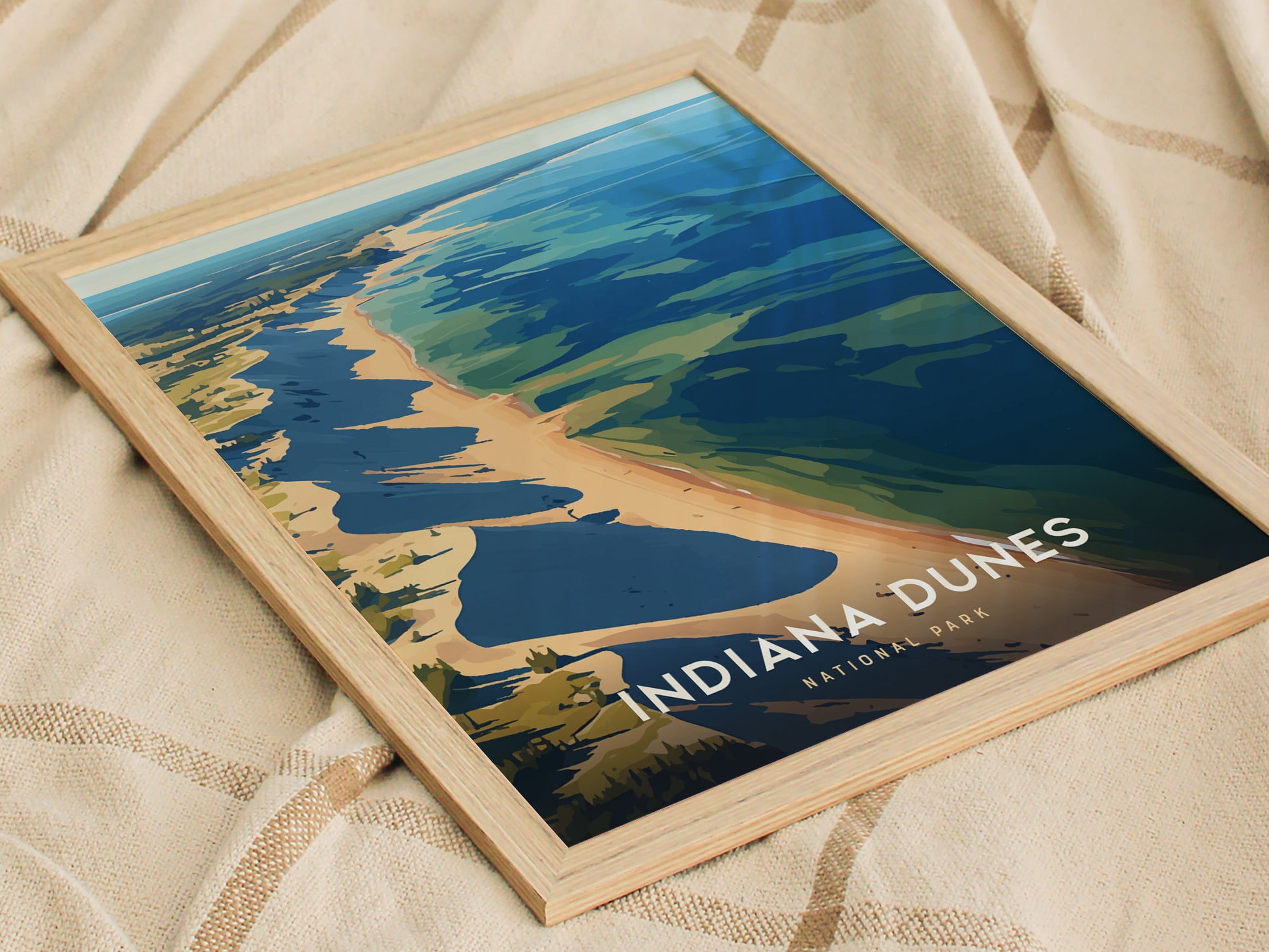 A framed poster of Indiana Dunes National Park lies on a beige fabric, depicting the shoreline and dunes with vibrant blue.
