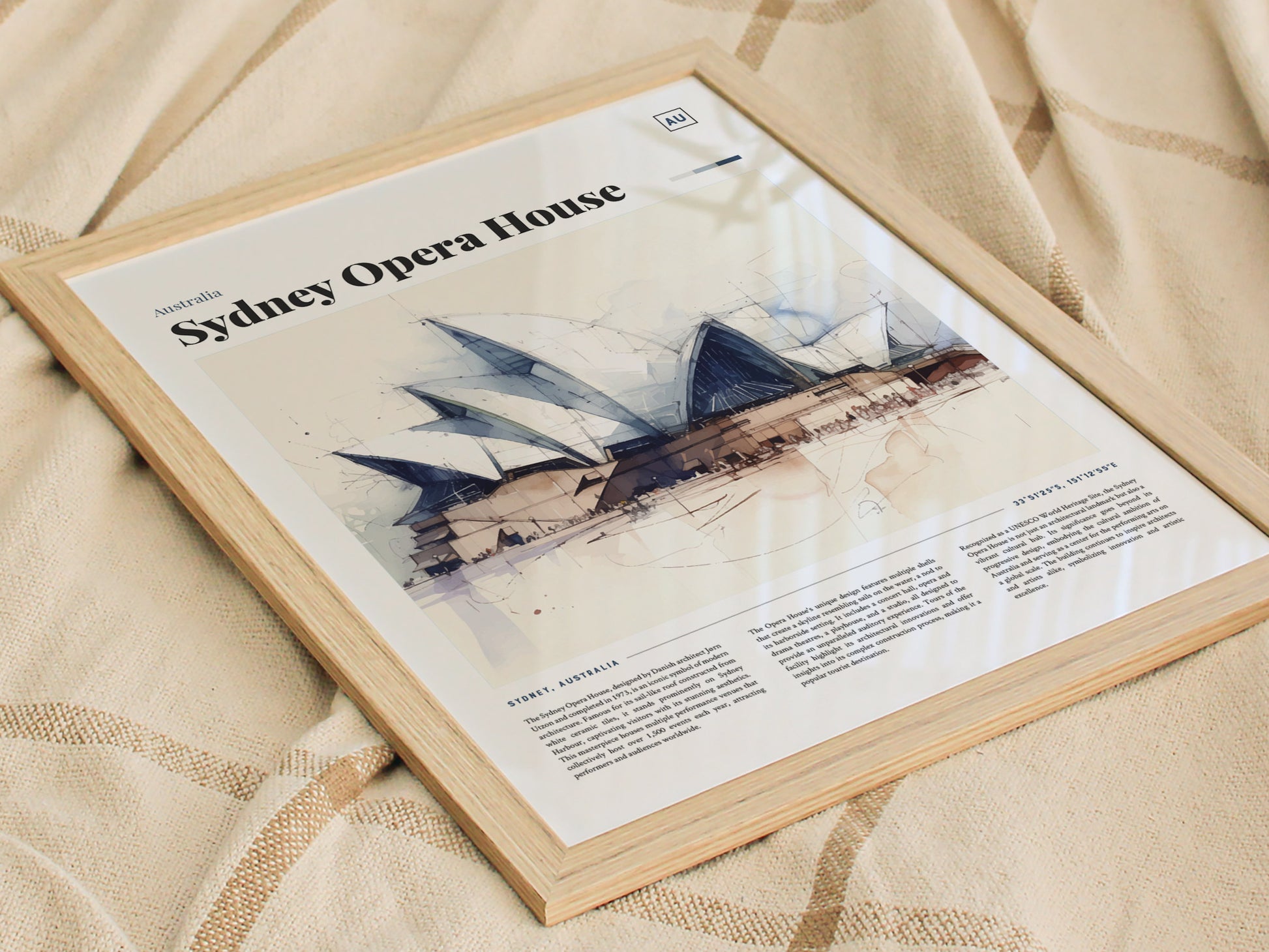 Framed poster of the Sydney Opera House featuring a watercolor illustration, resting on a beige patterned fabric. The image highlights the iconic sail-shaped architecture with descriptive text below.