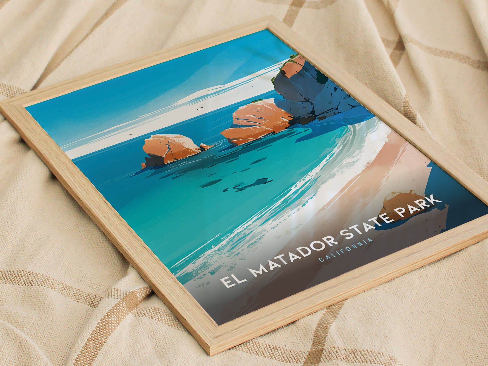 A framed poster of El Matador State Park, featuring vibrant blue ocean waves and rocky cliffs, rests on a beige fabric background. The text reads El Matador State Park, California.