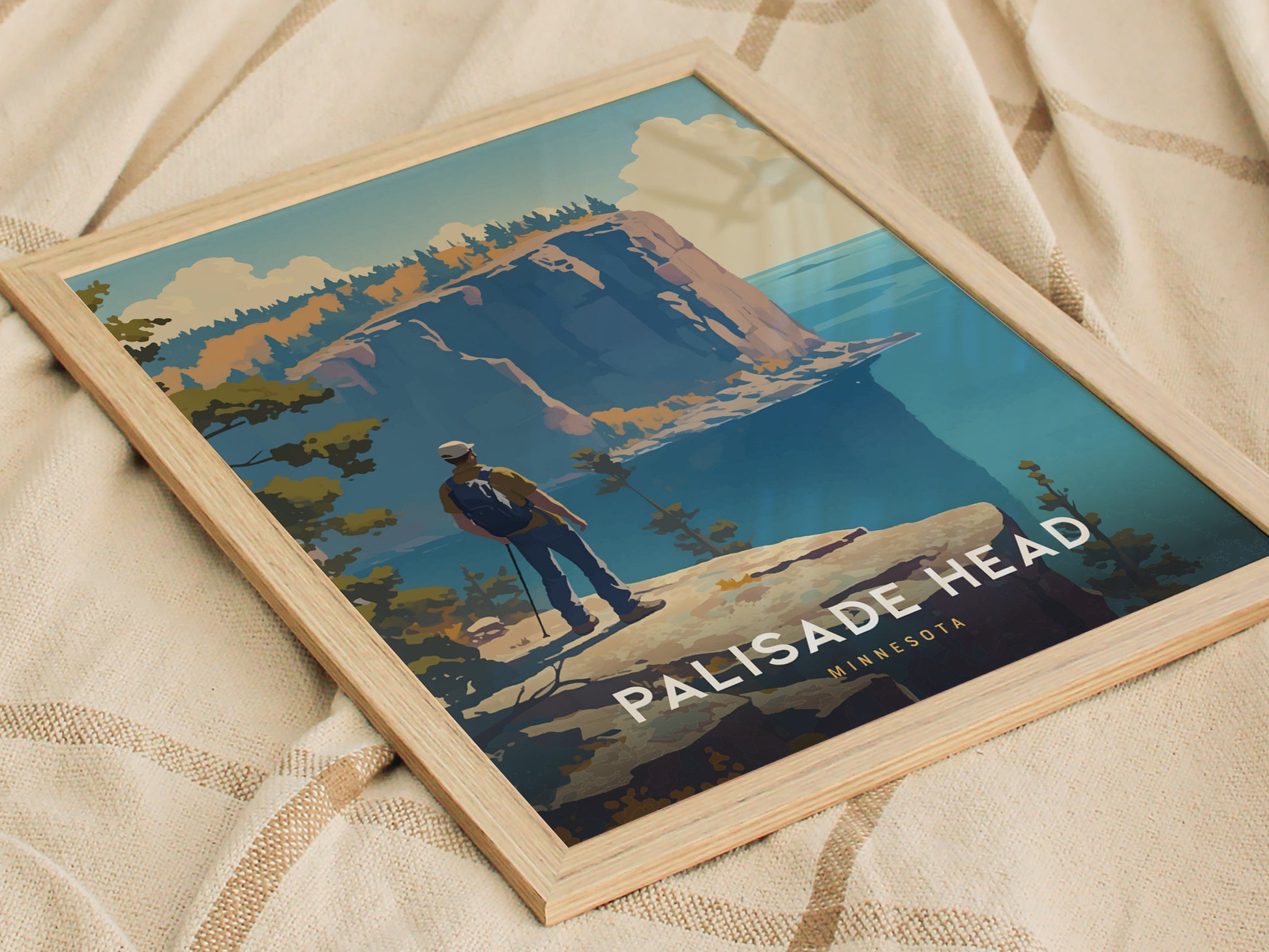 Framed Palisade Head Minnesota poster resting on beige fabric, featuring a hiker overlooking dramatic cliffs and Lake Superior, with vibrant trees and a clear blue sky.