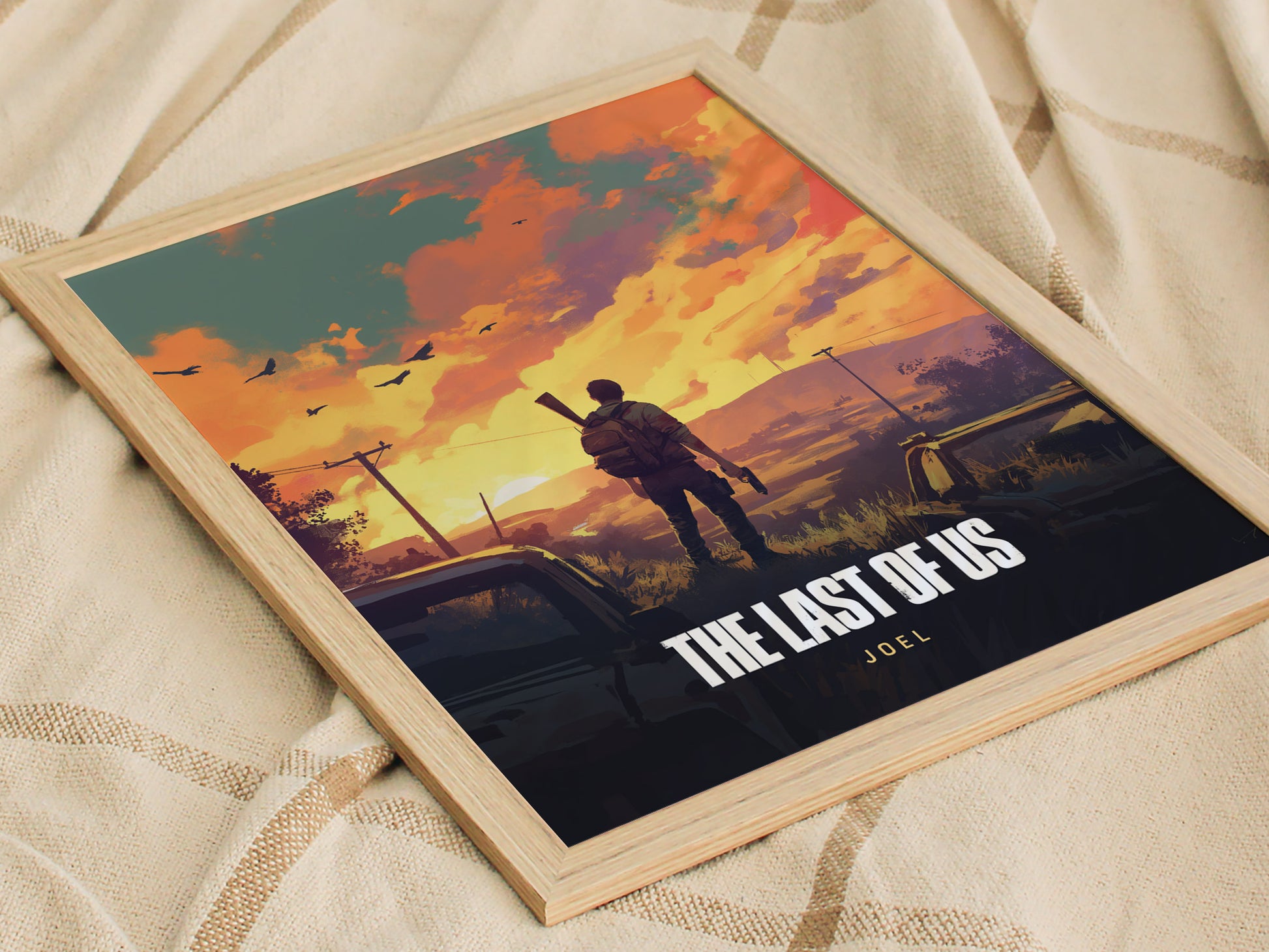 A framed poster on a fabric surface depicts a silhouette of a man with a gun against a vibrant sunset sky, featuring the text The Last of Us: Joel.