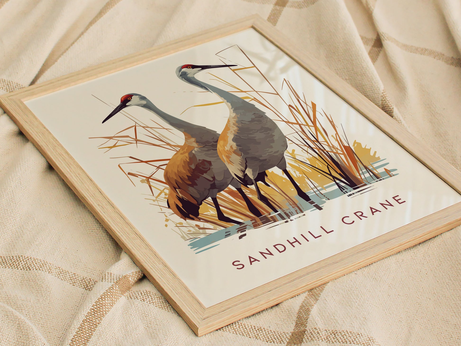 A framed poster displays two sandhill cranes standing amidst stylized marsh grasses with the text SANDHILL CRANE beneath, resting on a textured beige fabric.